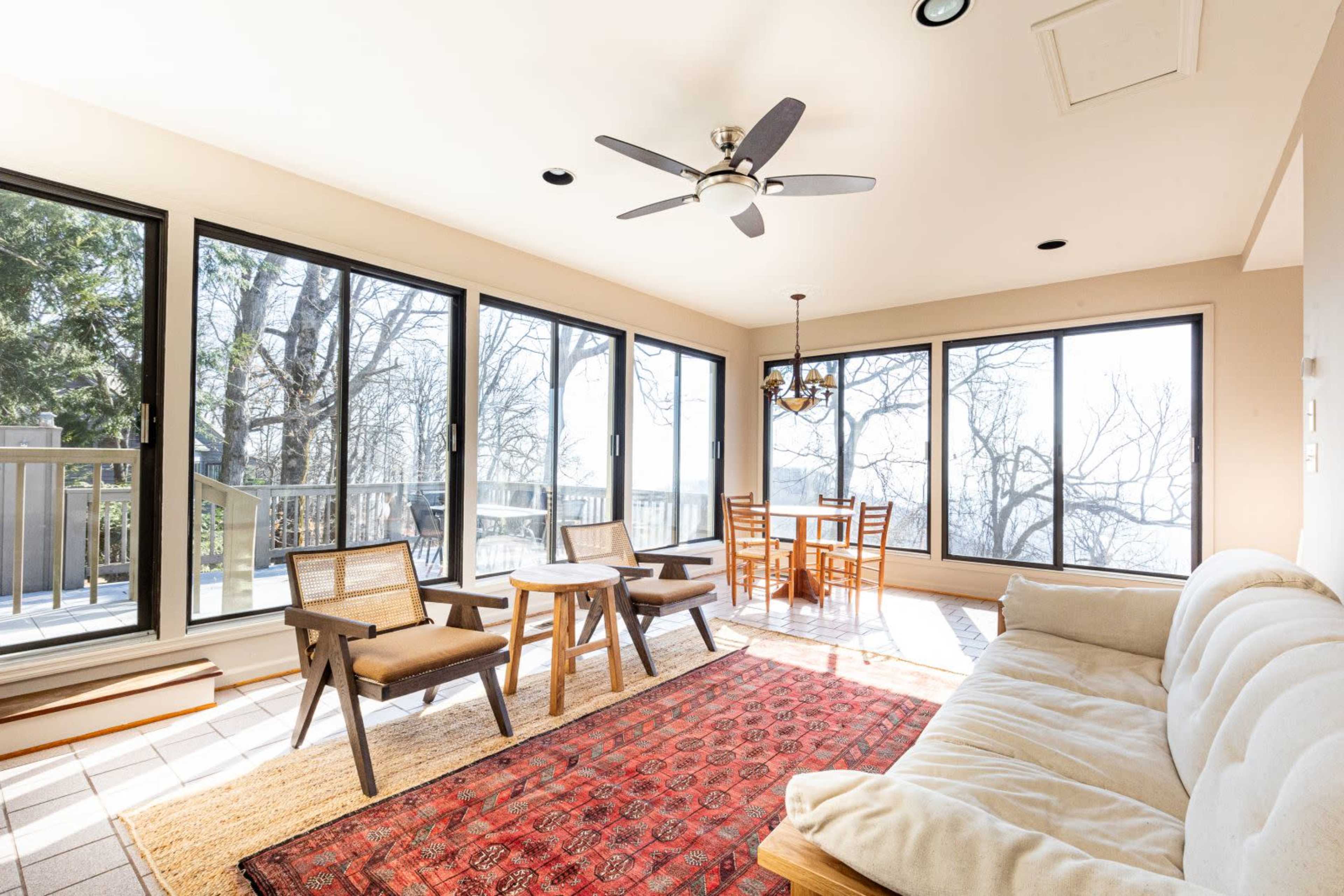 A sunlit sunroom with large windows overlooking trees, featuring a seating area with a sofa, two chairs, a table, and a chandelier.
