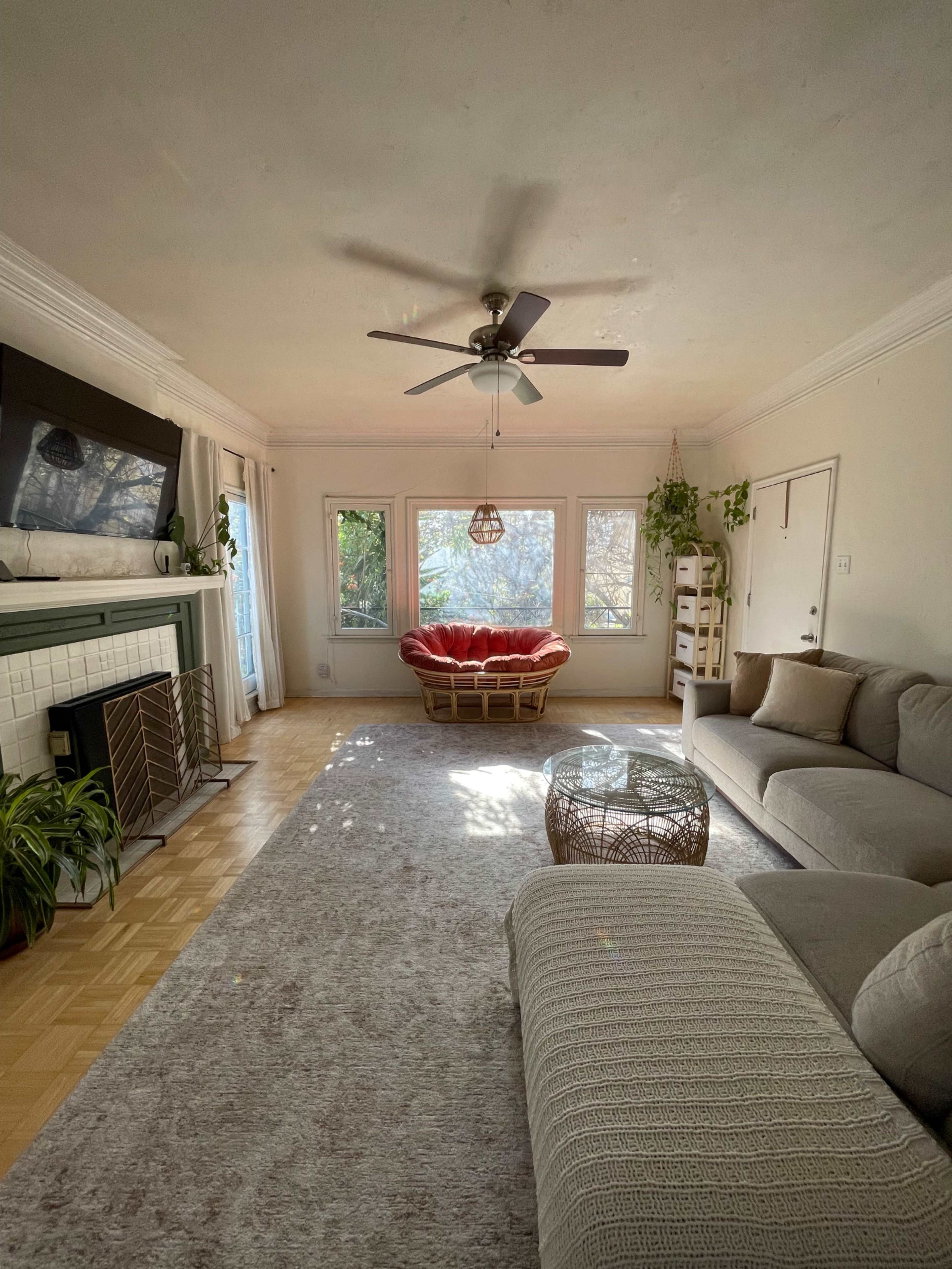A spacious living room features a light-colored sofa, a circular coffee table, a large window with greenery outside, and a cozy chair in the corner.