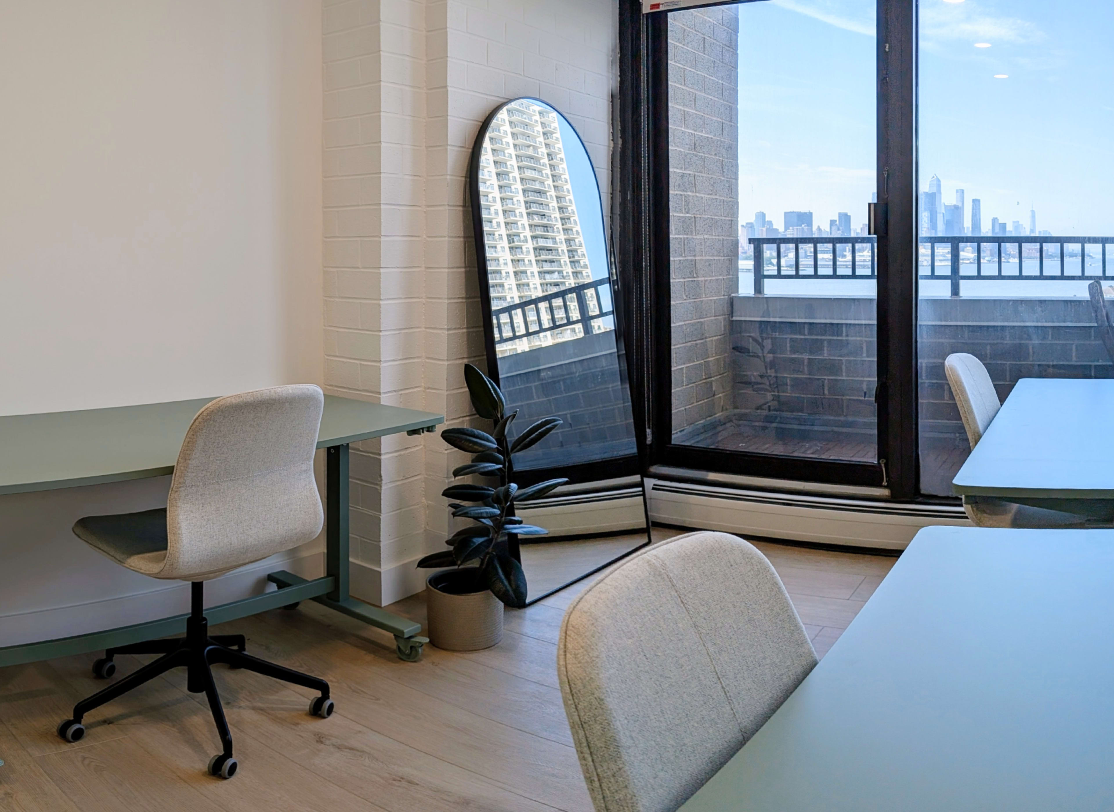 The image shows a bright office space featuring two light green desks with grey chairs, a large mirror reflecting a city skyline, and a potted plant in the corner.