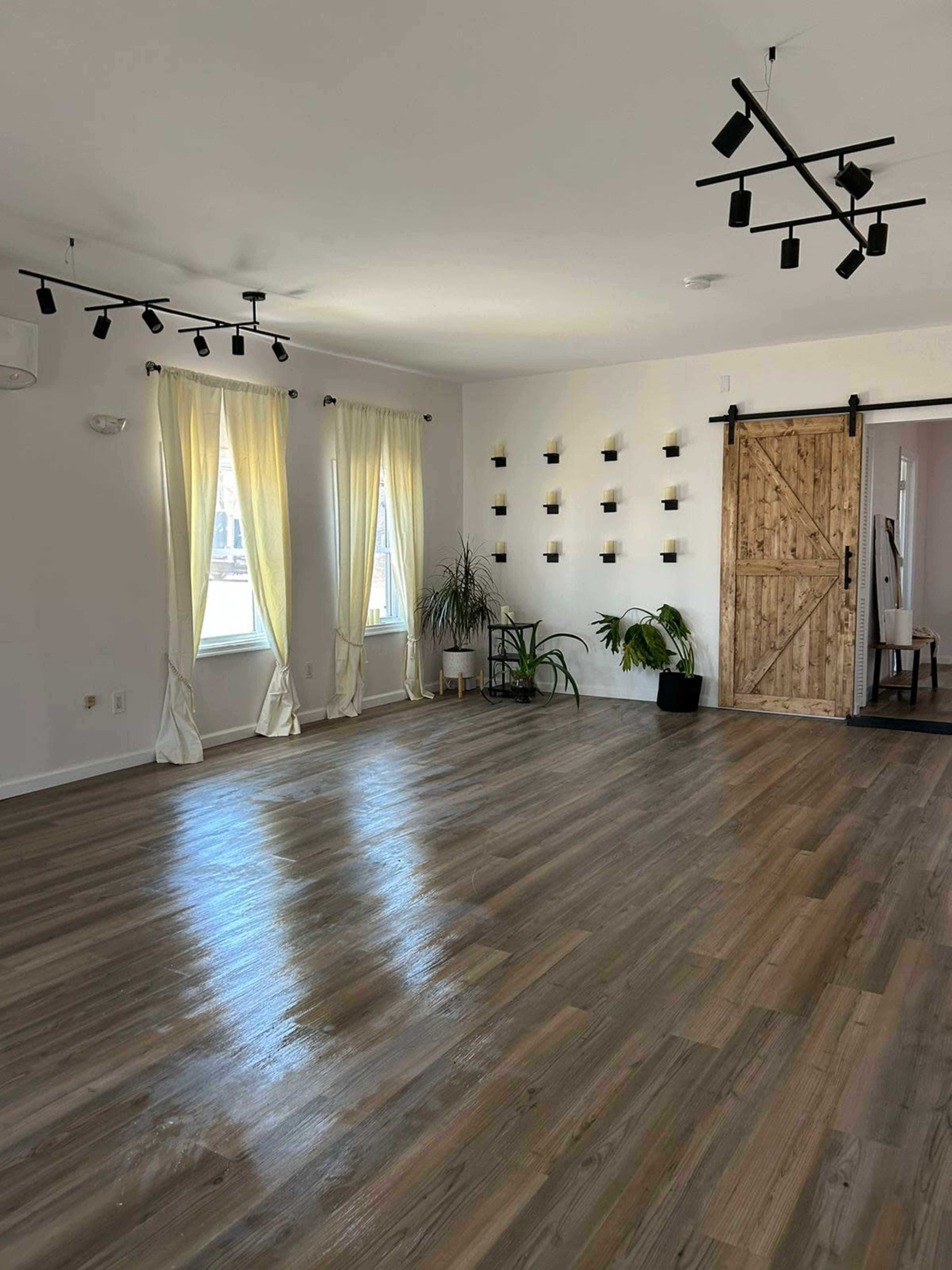 A spacious, well-lit room with wooden flooring, large windows draped with light curtains, and a wooden sliding door.