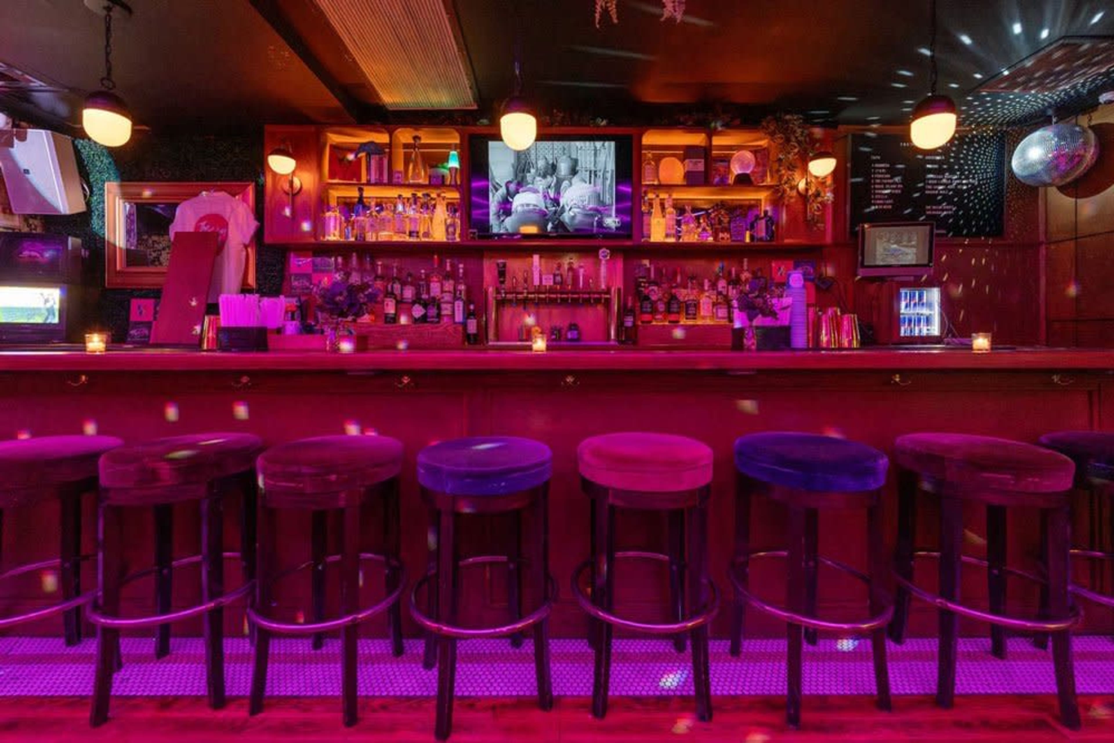 The image shows a dimly lit bar with a long counter, high stools, and shelves filled with various bottles, illuminated by colorful lights.