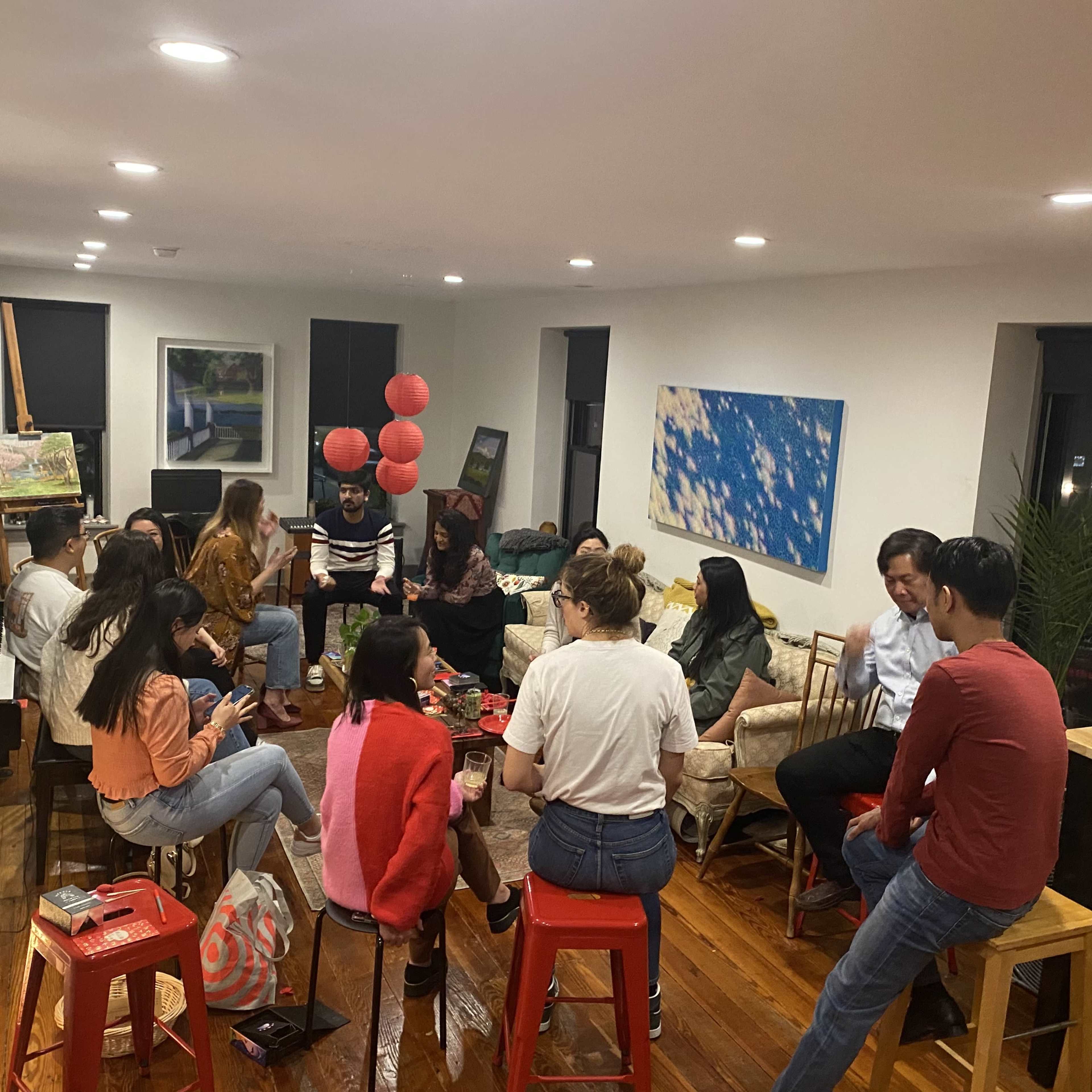 A group of people is seated in a well-lit living room, arranged in a circle around a central coffee table adorned with snacks and drinks.