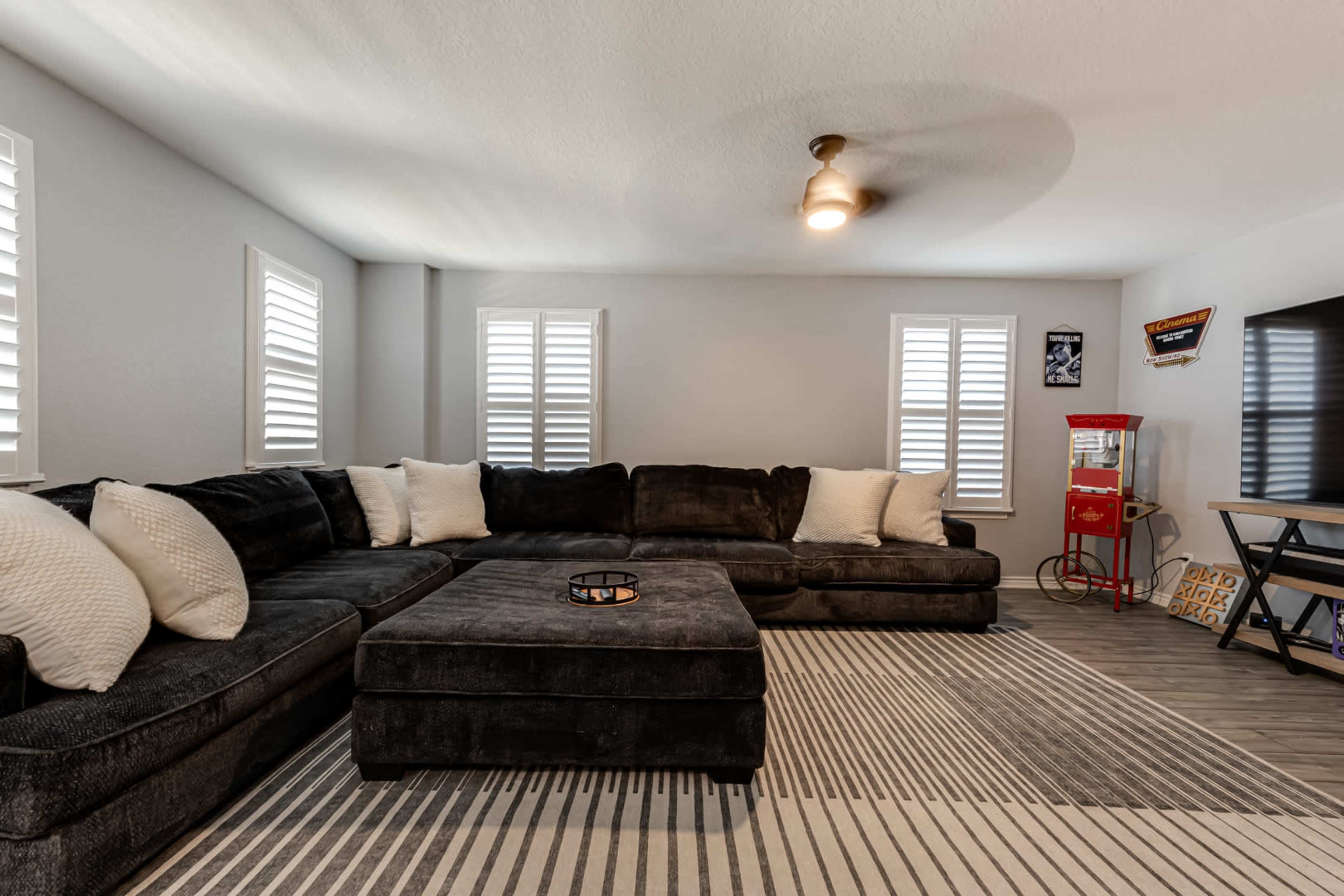 A modern living room featuring a large black sectional sofa, a coffee table, and a popcorn machine, with light streaming through shuttered windows.