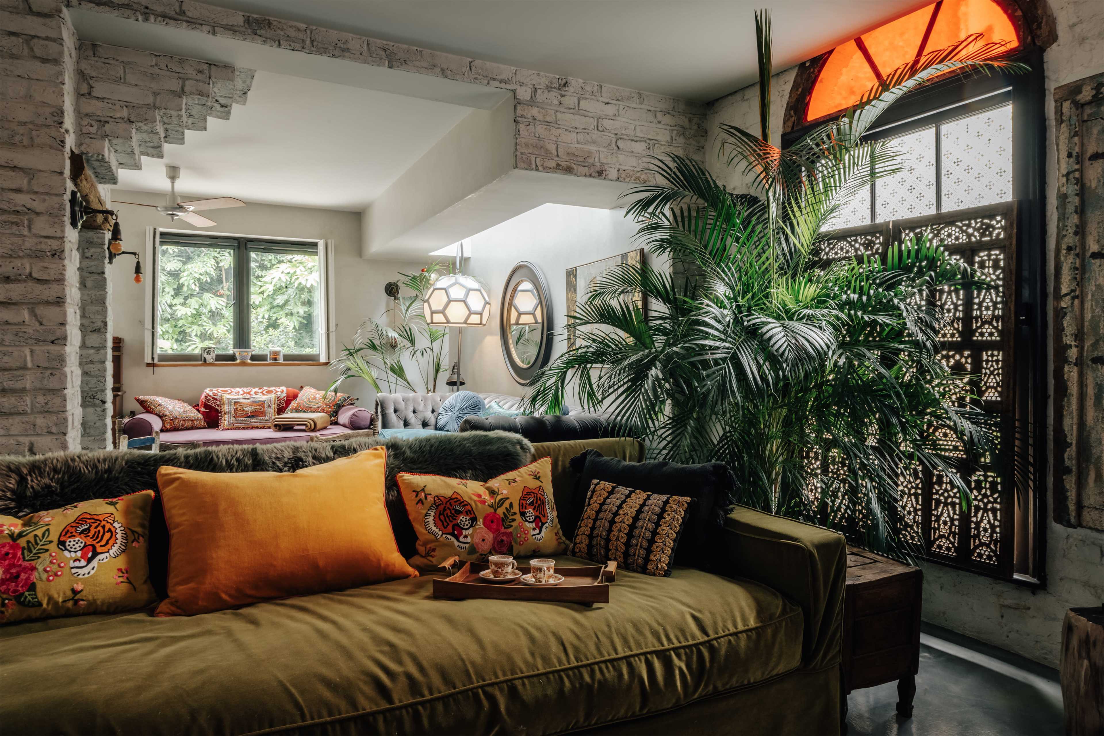 A spacious living room features a green couch adorned with colorful cushions and a wooden tray, surrounded by large plants and bathed in natural light.