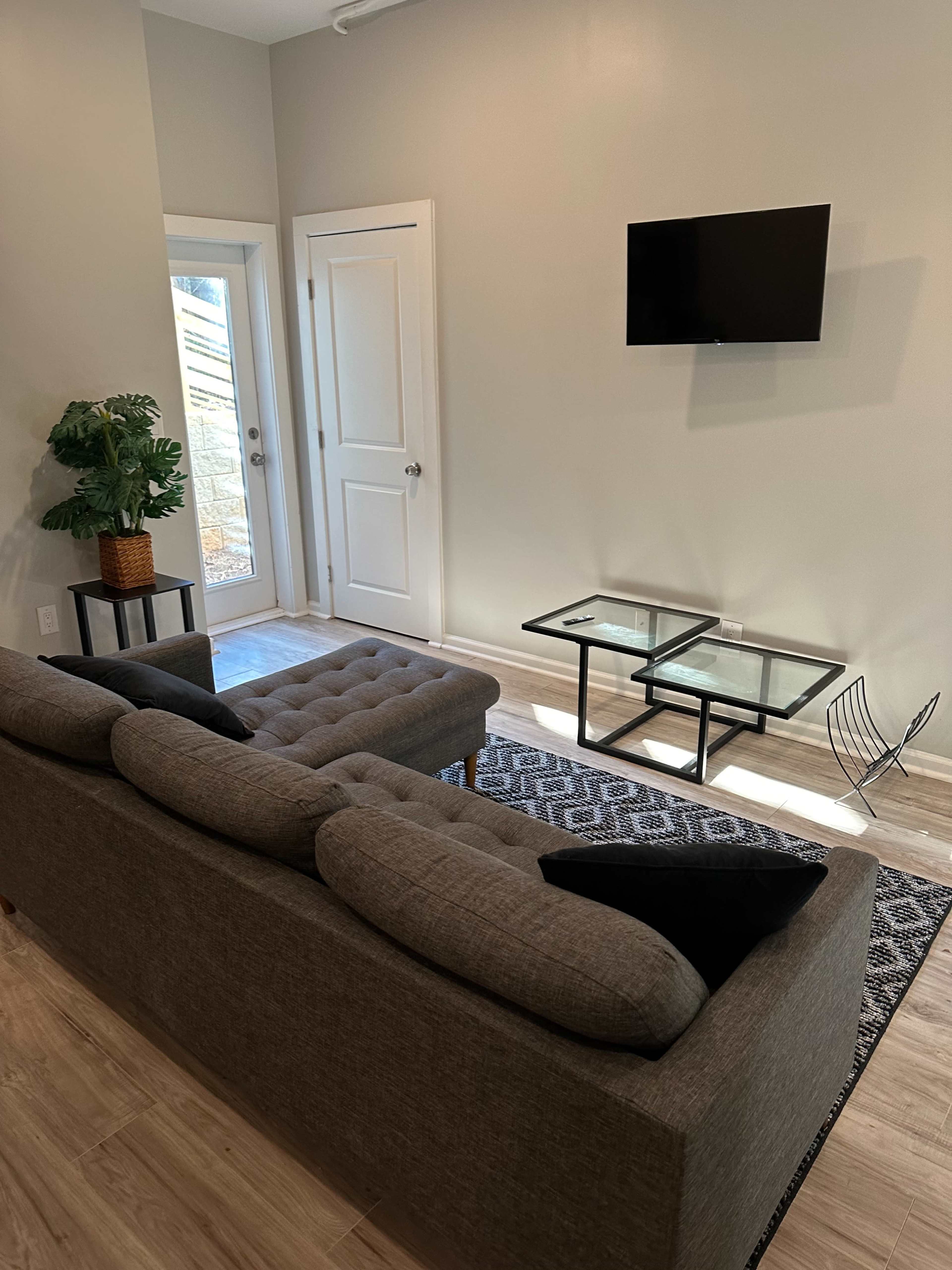 The image shows a living room with a gray sectional sofa, a glass coffee table, a small plant in a basket, and a wall-mounted television.