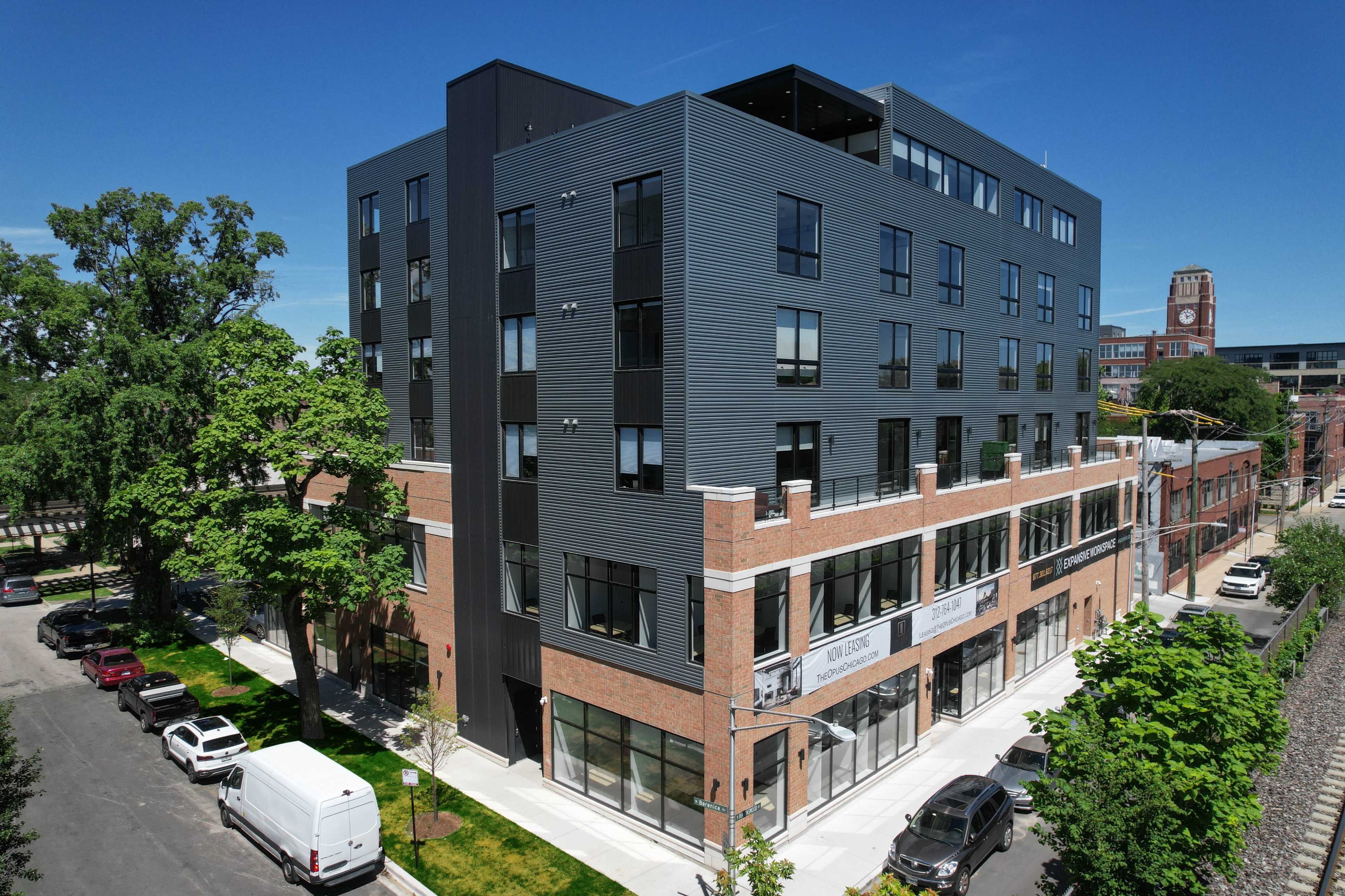 The image shows a modern five-story building with a combination of brick and metal siding, located on a tree-lined street.