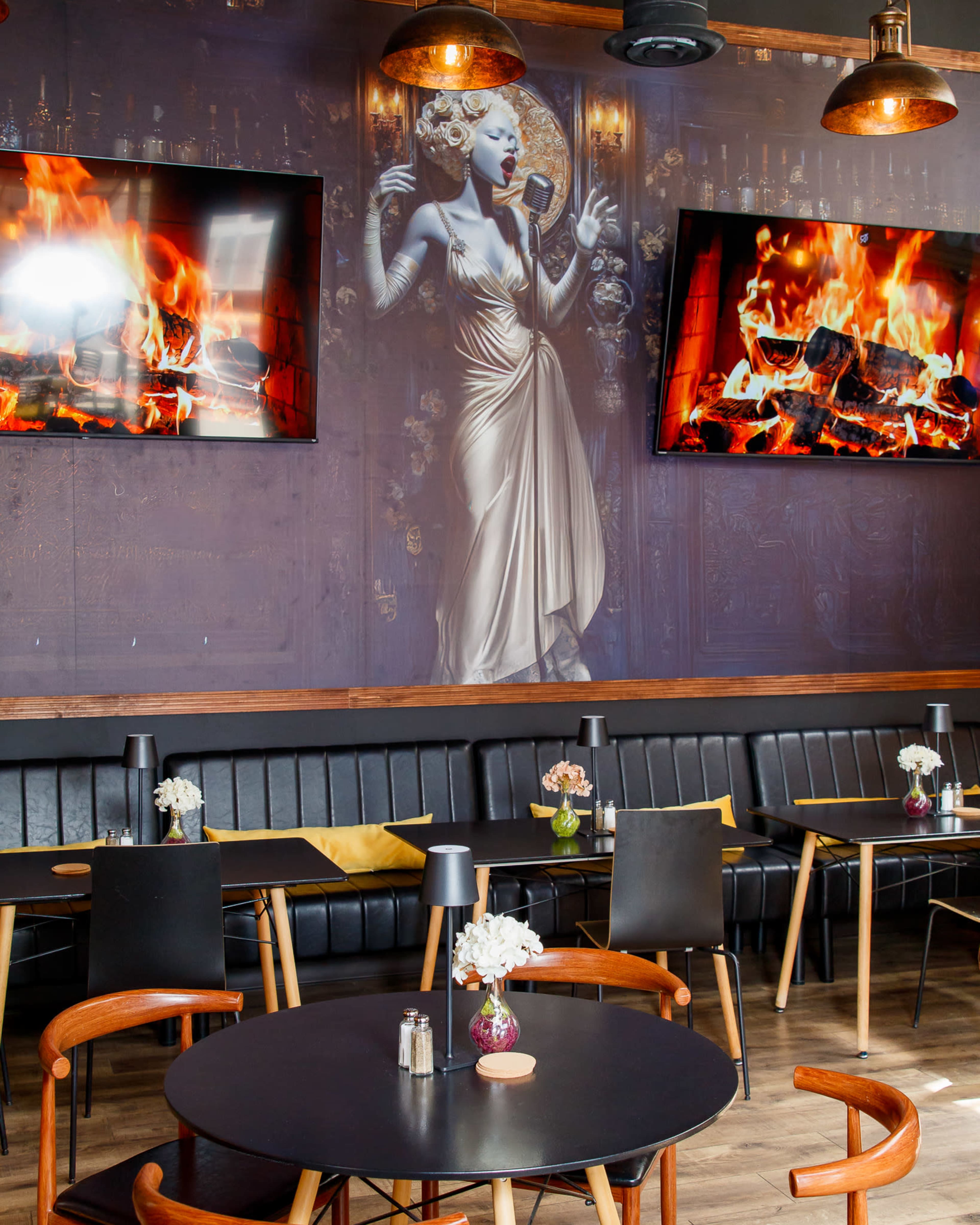 The image shows a stylish restaurant interior featuring a large mural of a woman singing, paired with two screens displaying flames, and black booths with round tables adorned with small flower arrangements.
