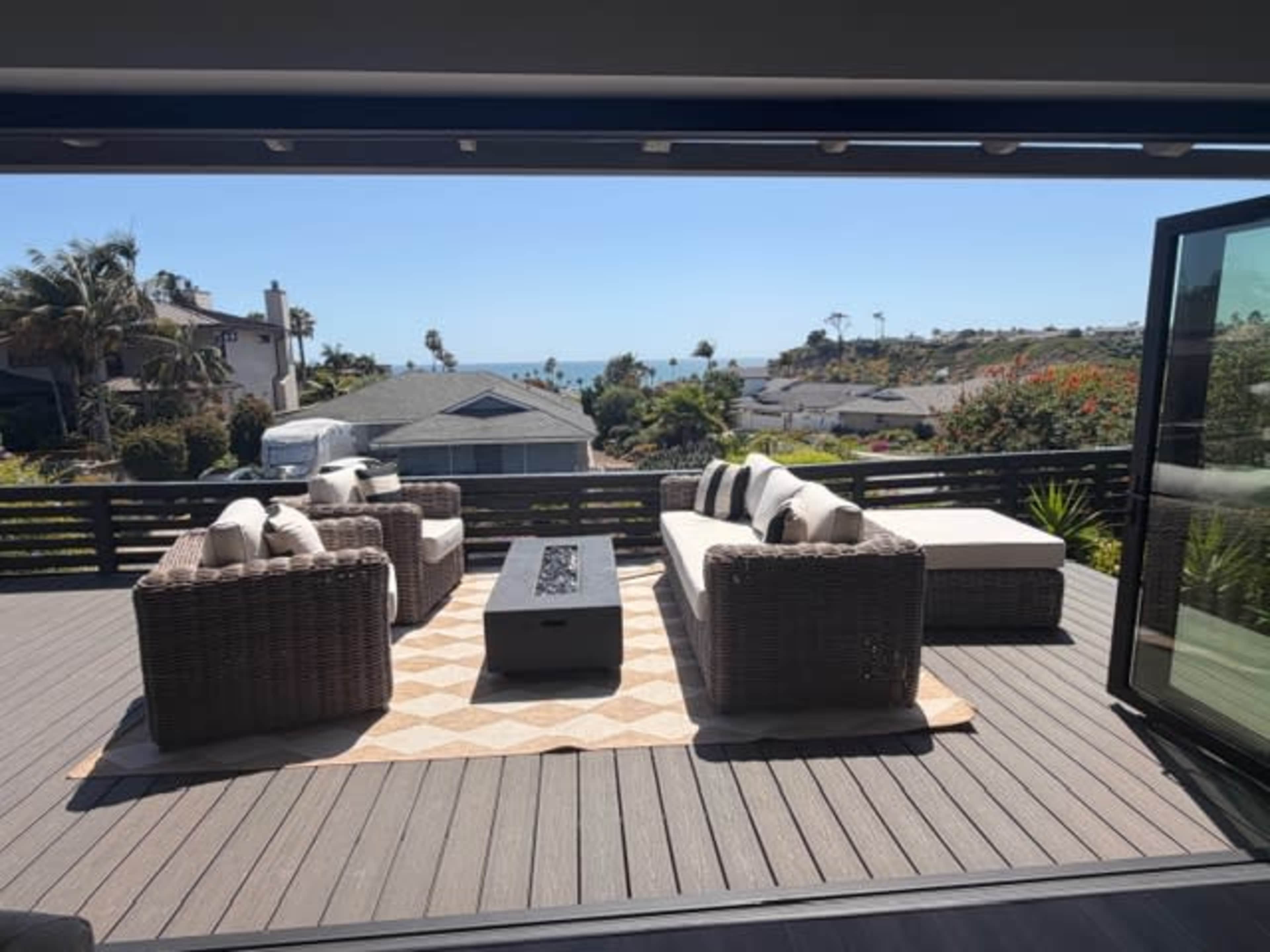 A patio overlooking a coastal view features outdoor furniture arranged around a fire pit on a wooden deck.