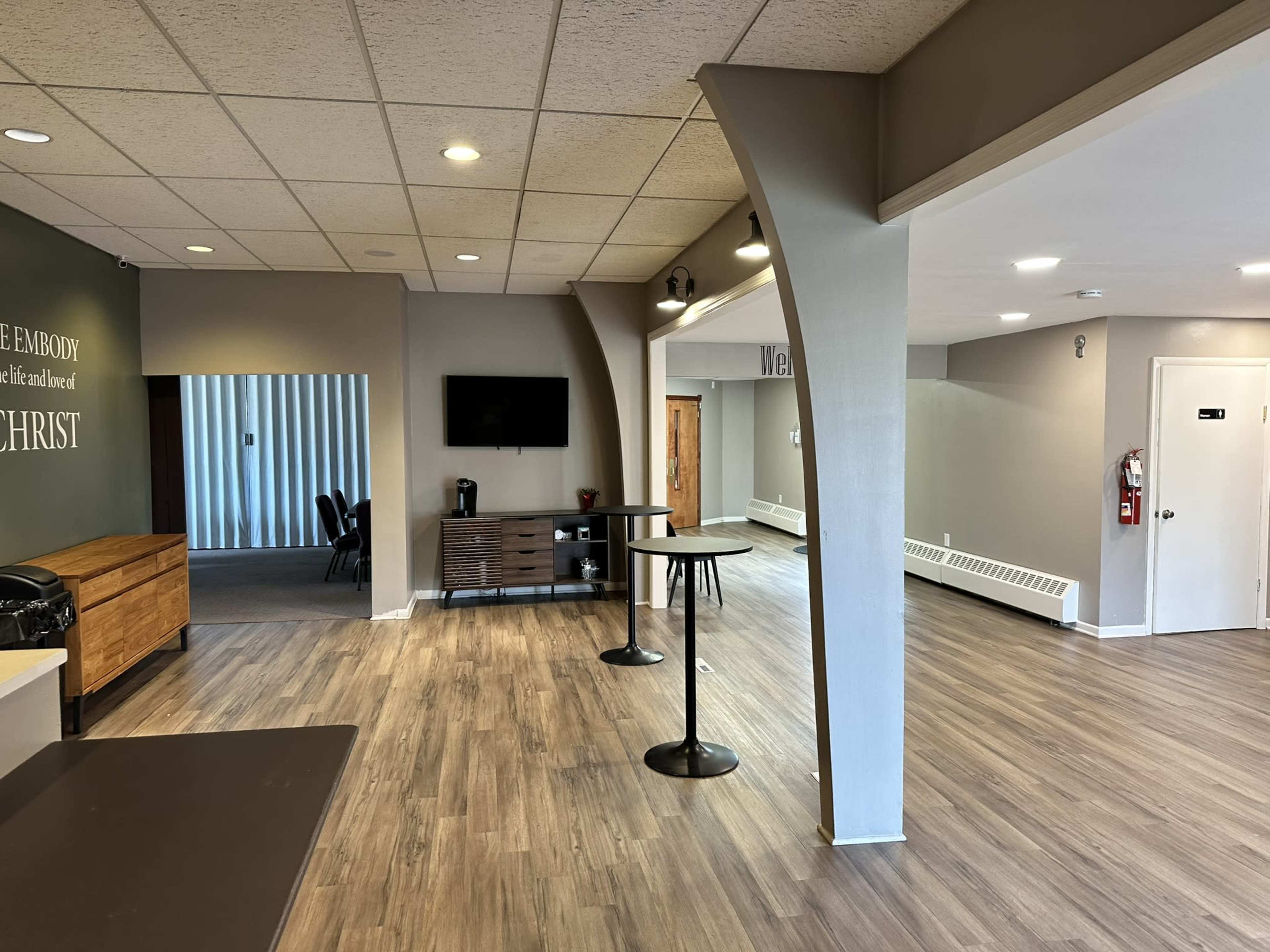 A spacious interior area with wooden floors, showcasing modern furniture, a television, and a reception desk against a light gray wall.
