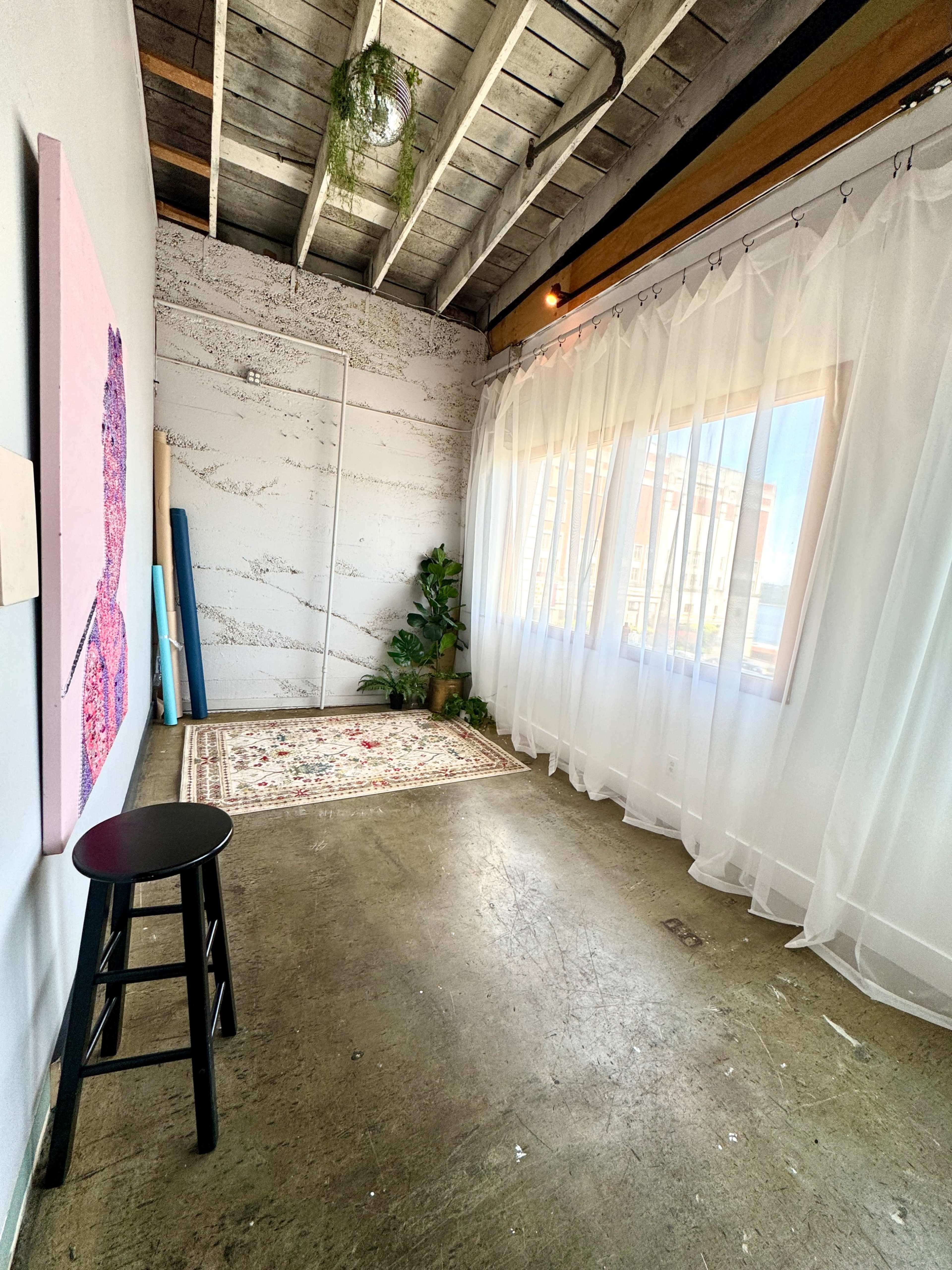 The image shows a spacious, well-lit room with a wooden ceiling, a large window covered by sheer curtains, and minimal furnishings including a stool and a decorative rug.