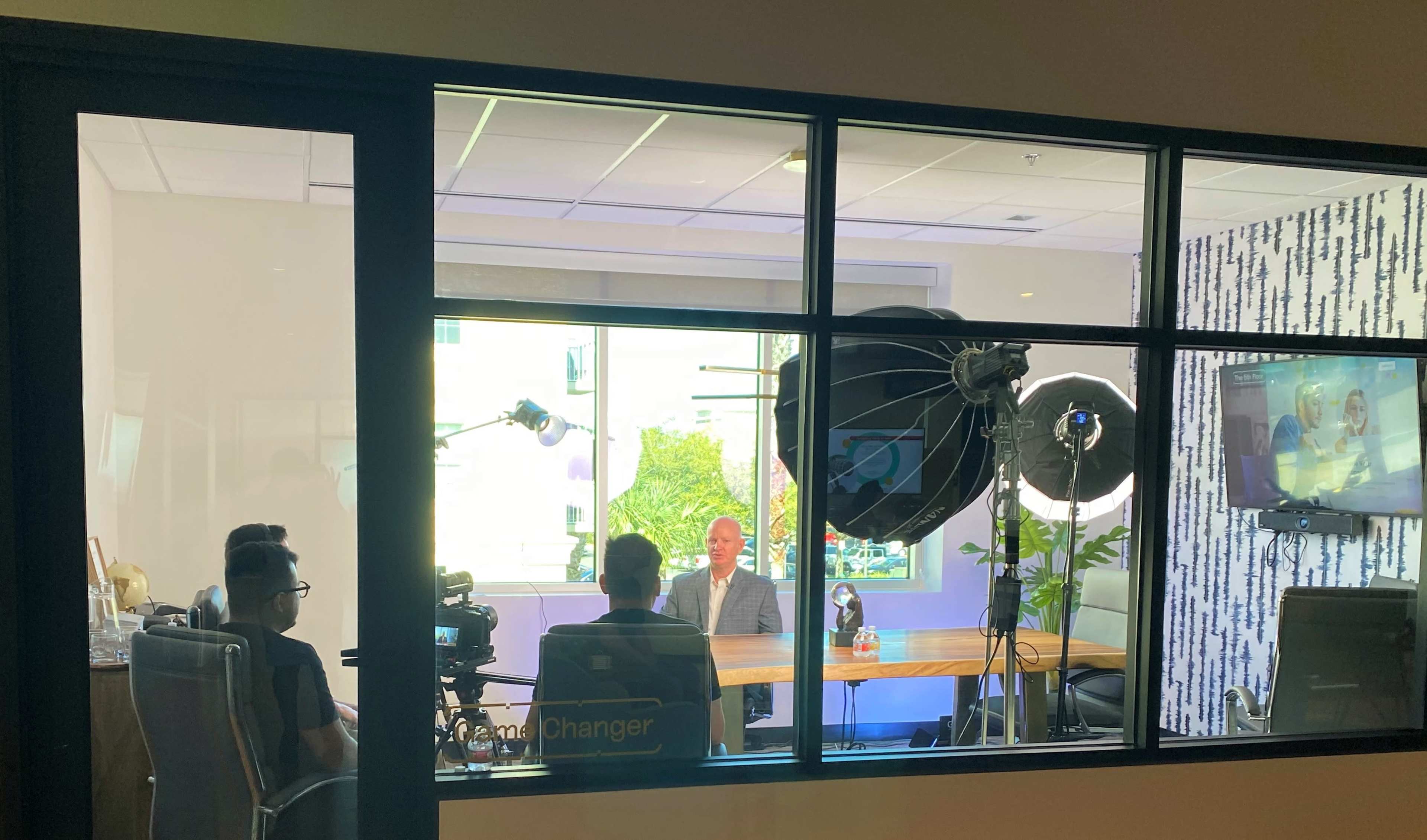 A group of people is filming an interview inside a well-lit office with large windows and studio equipment set up.
