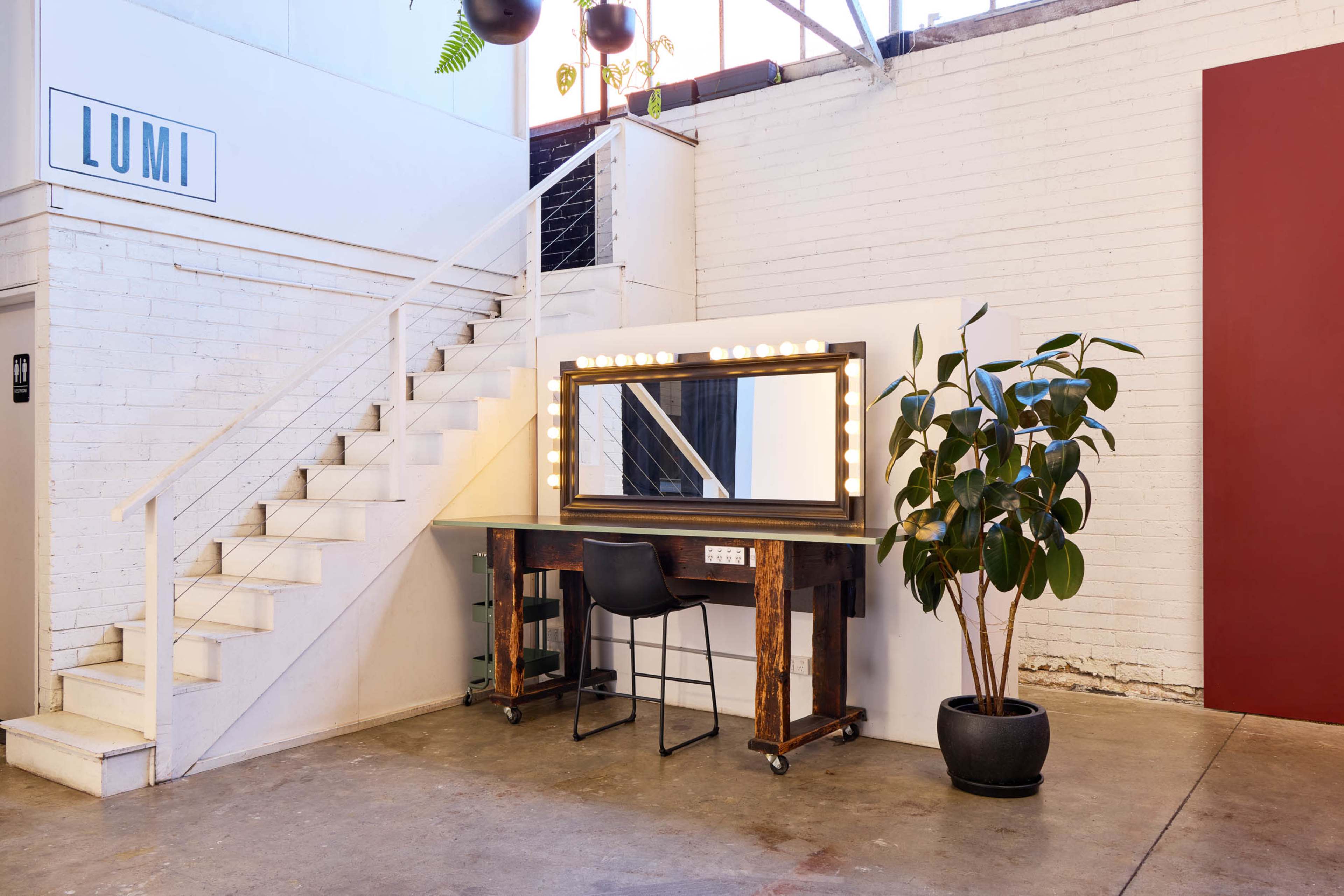The image shows a modern interior with a makeup station featuring a large mirror surrounded by lights, next to a stairway and a potted plant.