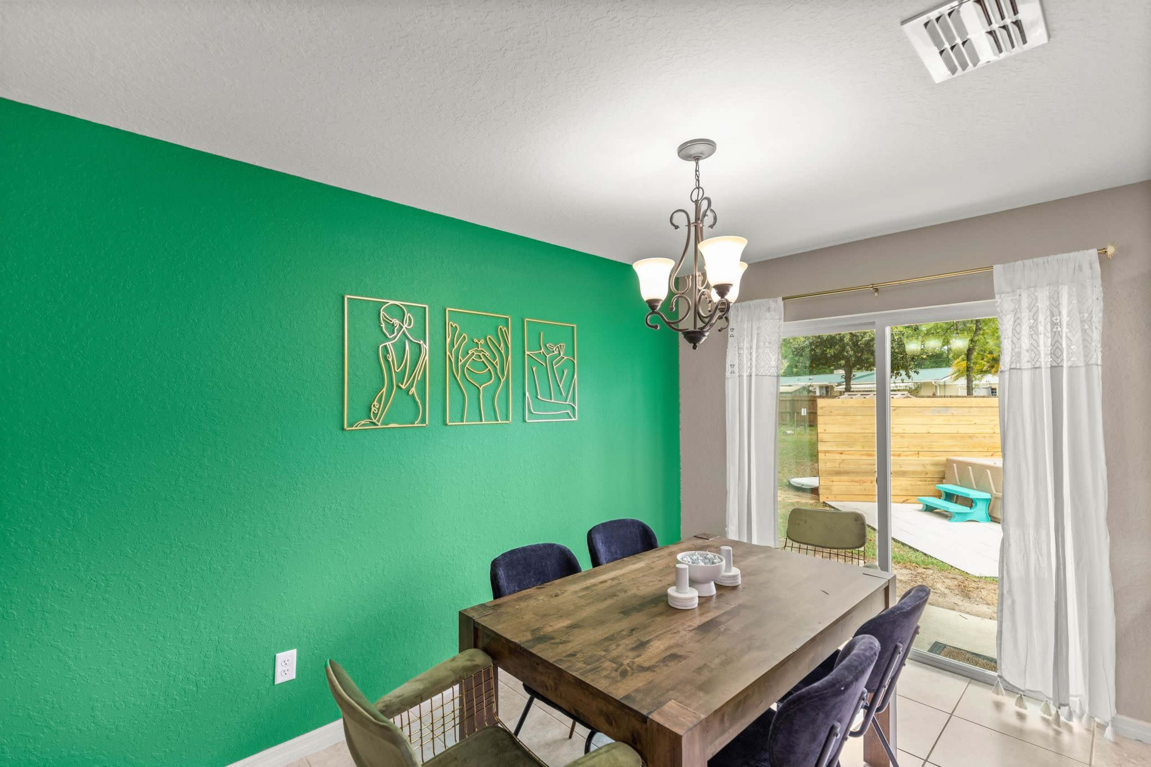A dining area features a wooden table surrounded by chairs, with green walls and artistic wall hangings, and a sliding door leading to an outdoor space.