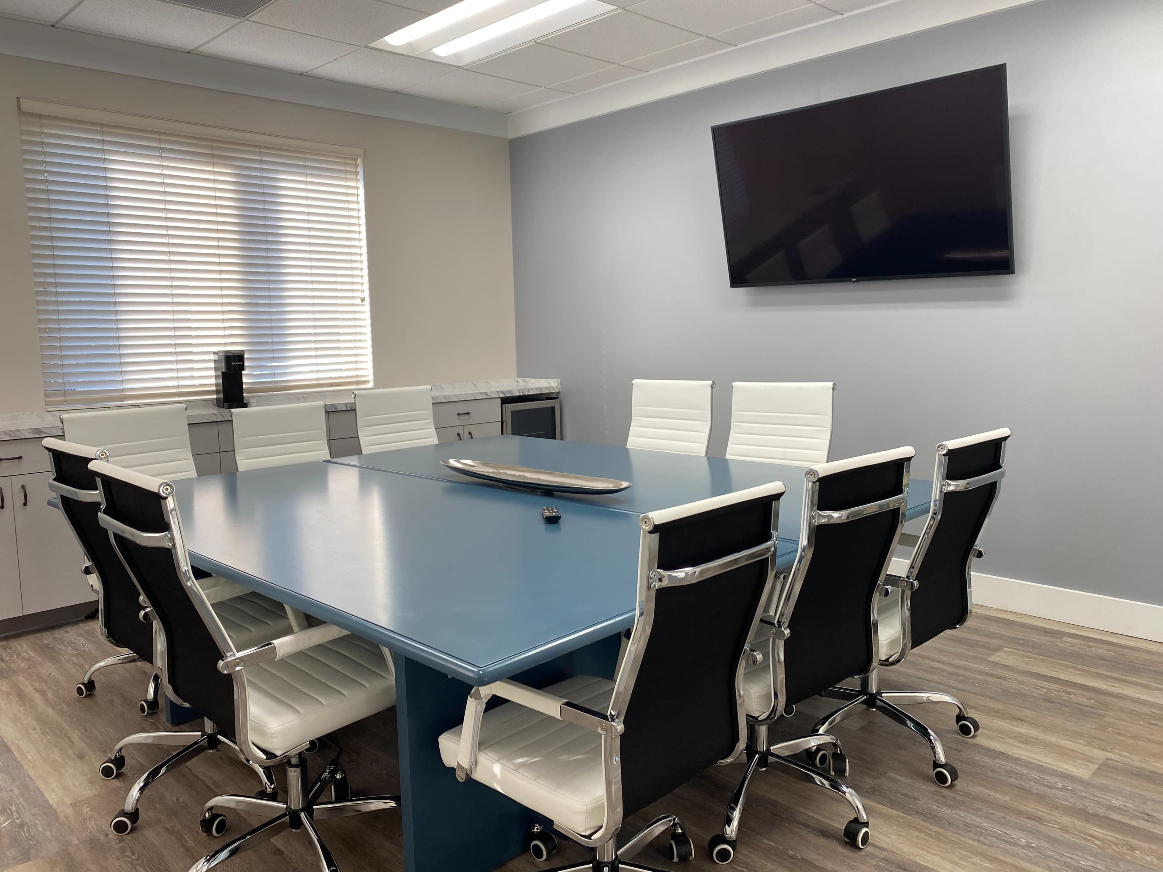 A modern conference room with a large blue table surrounded by white and black chairs, a wall-mounted TV, and a window with blinds.