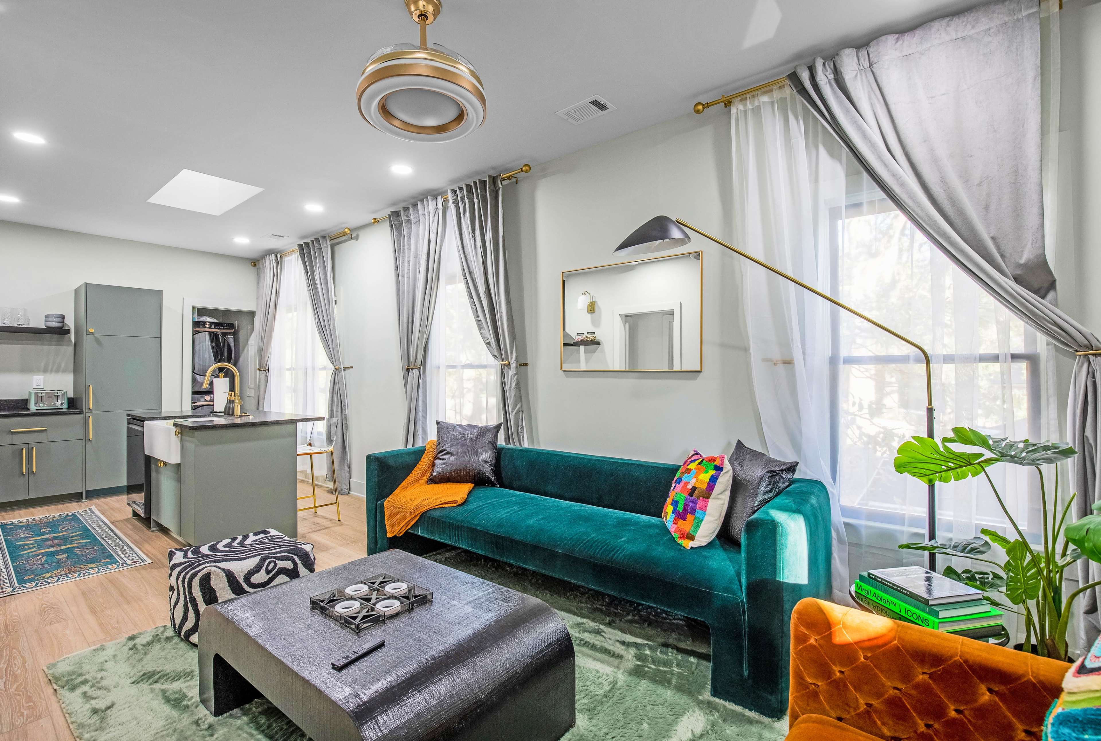 A modern living room with a green velvet sofa, a black coffee table, and a kitchen area in the background, adorned with large windows and elegant curtains.