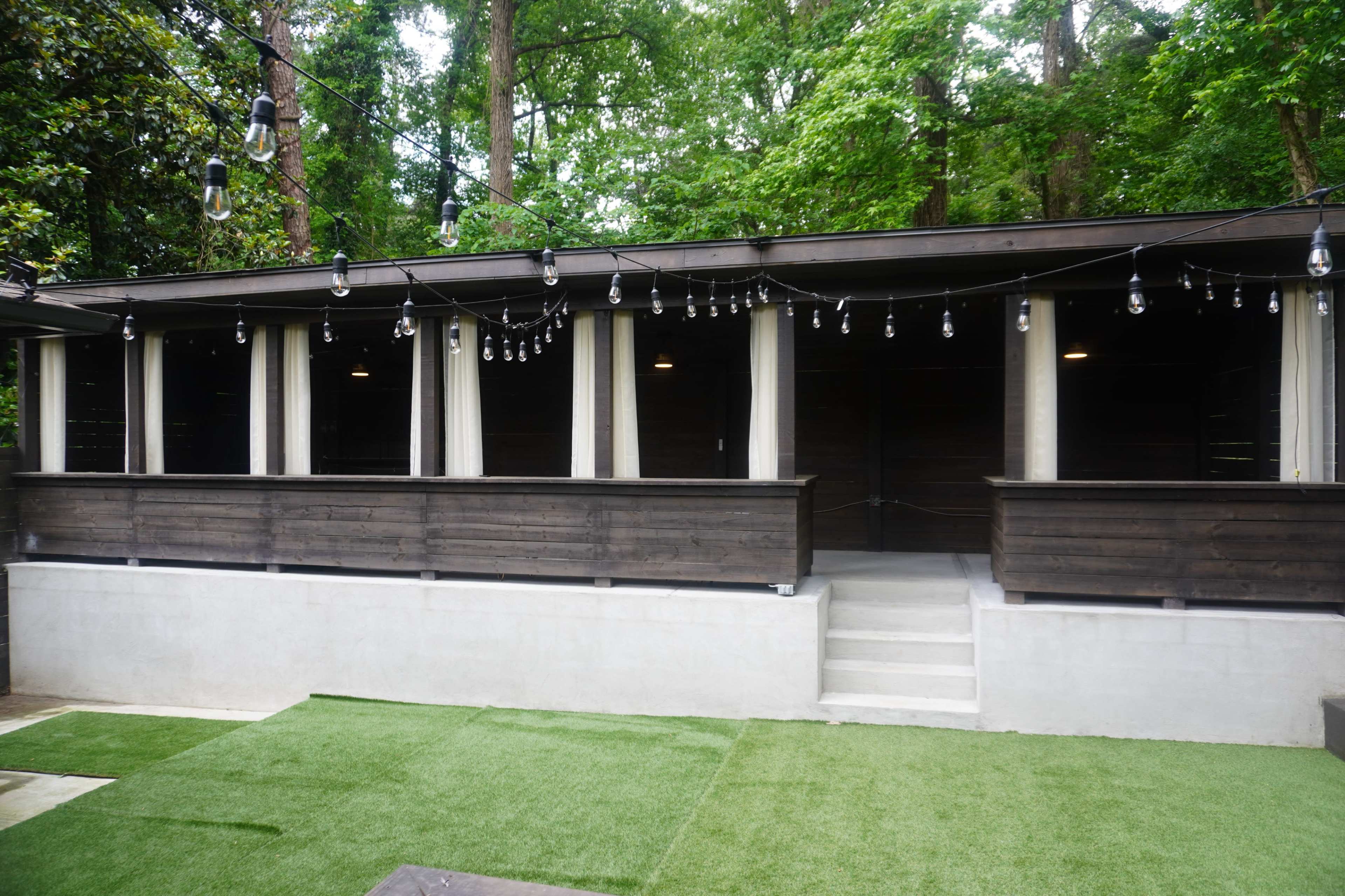 The image shows a modern wooden structure with multiple windows set against a backdrop of greenery, accompanied by string lights overhead.
