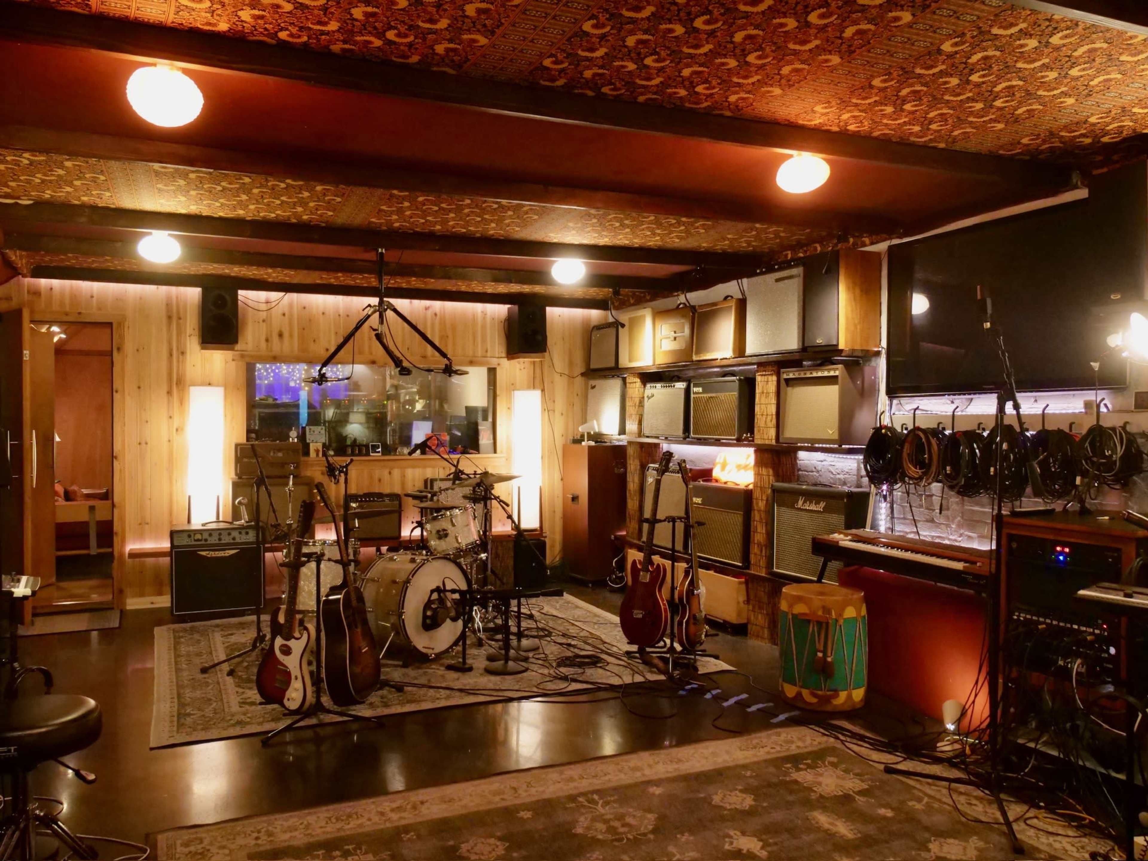 The image shows a music recording studio equipped with various musical instruments, amplifiers, and sound recording equipment arranged in a well-lit space with wooden and patterned ceilings.