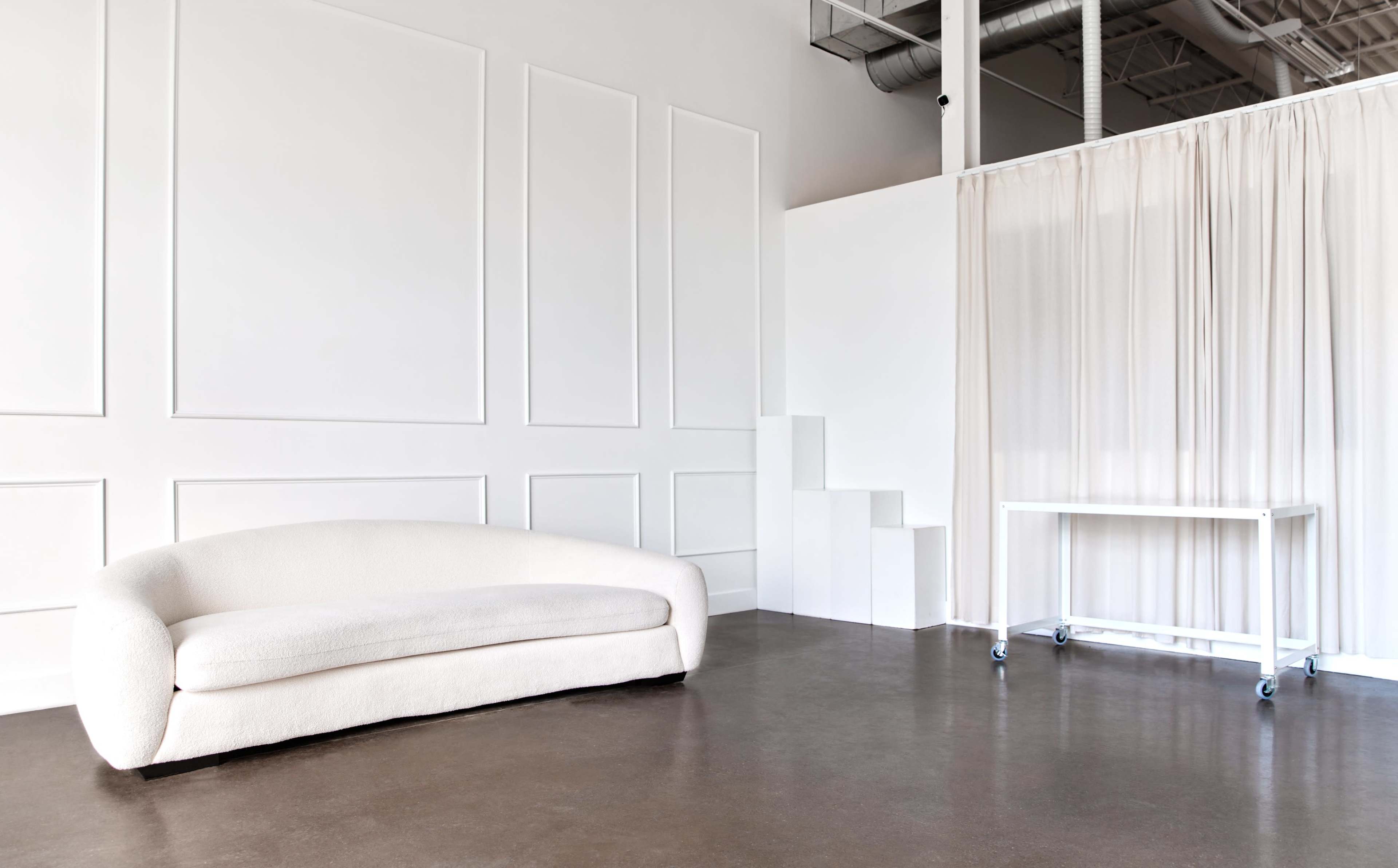 A minimalist interior featuring a white curved sofa, a plain white wall with paneling, and a transparent curtain dividing the space.