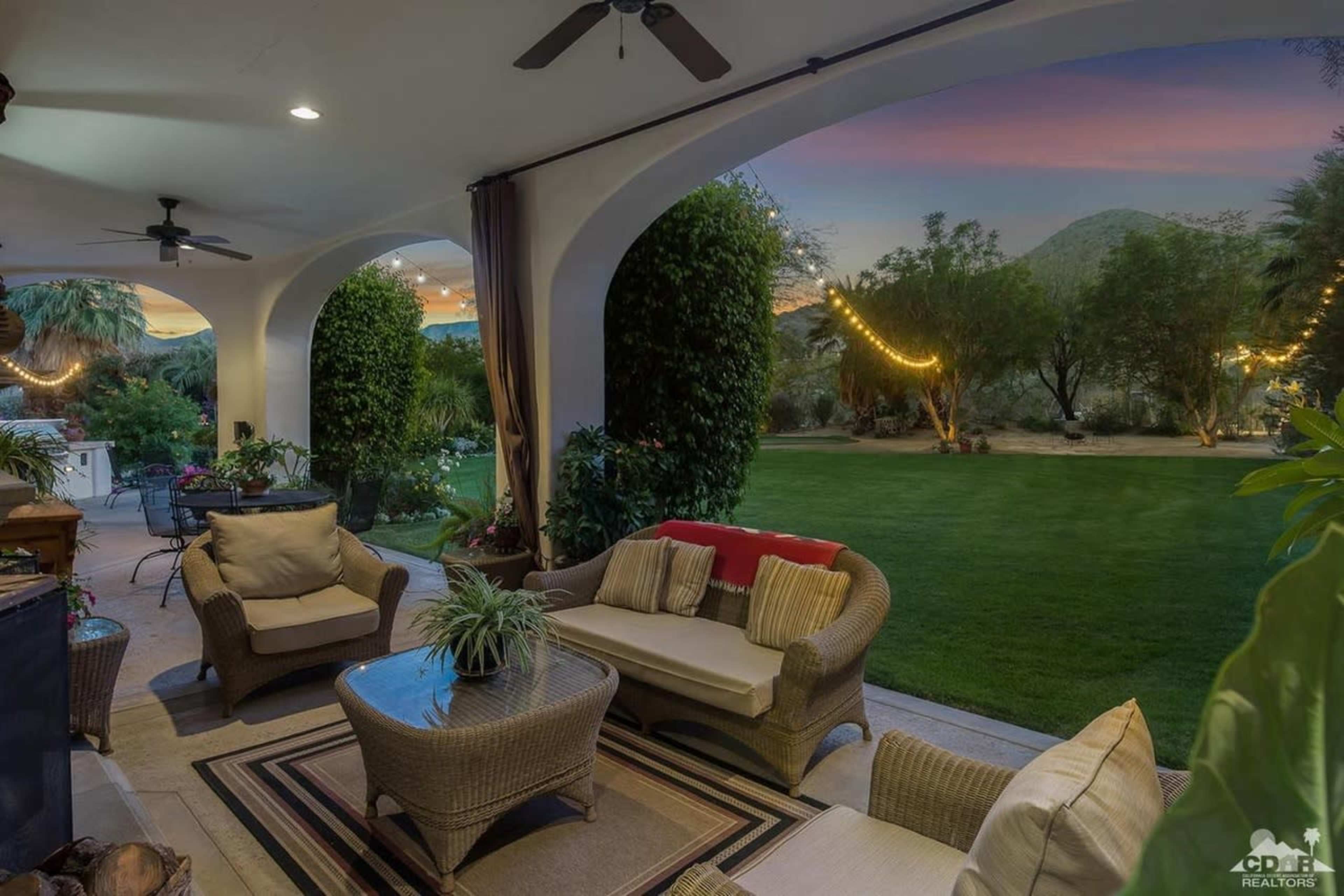 A spacious patio features comfortable seating and a view of a well-maintained lawn framed by trees, with hanging lights illuminating the scene at dusk.