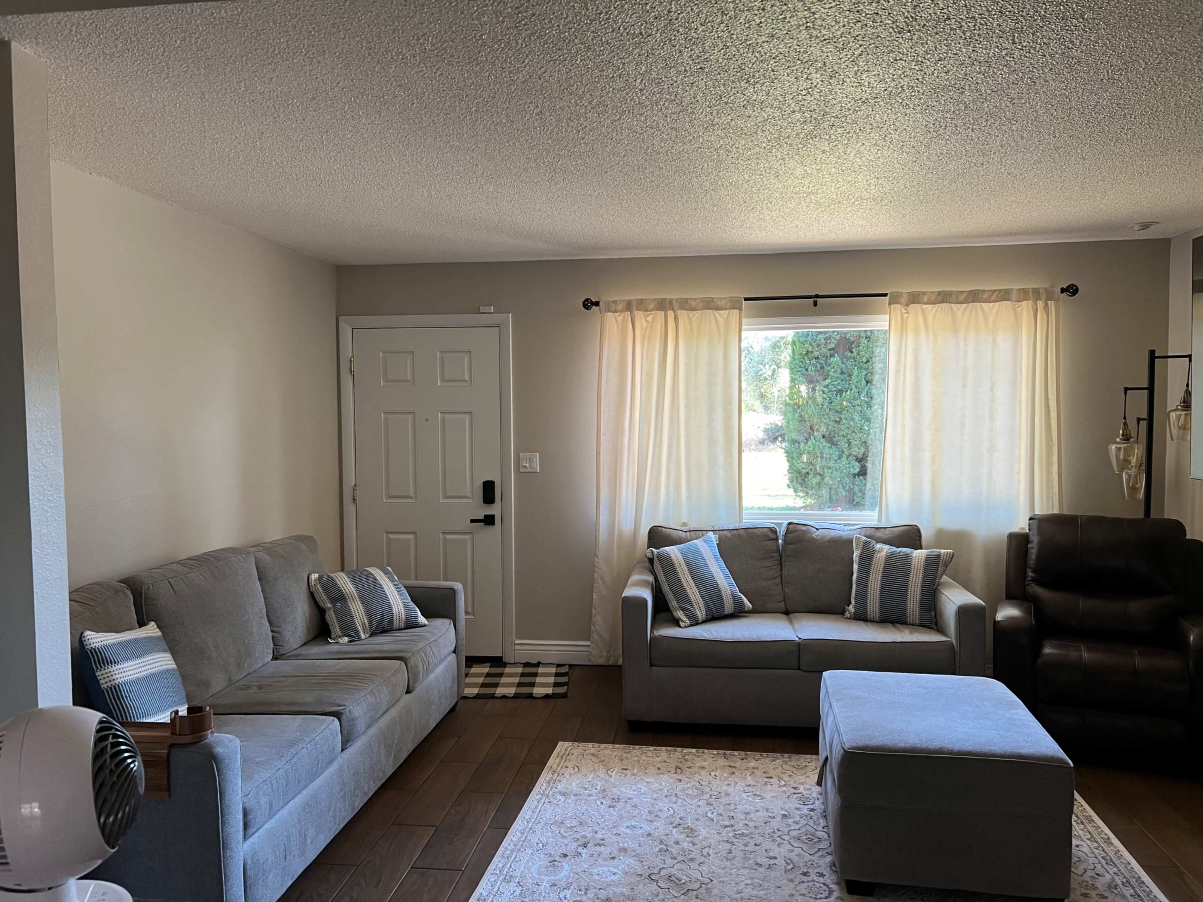 The image shows a living room with two gray sofas, a footstool, and a door leading outside, illuminated by natural light through a large window with sheer curtains.