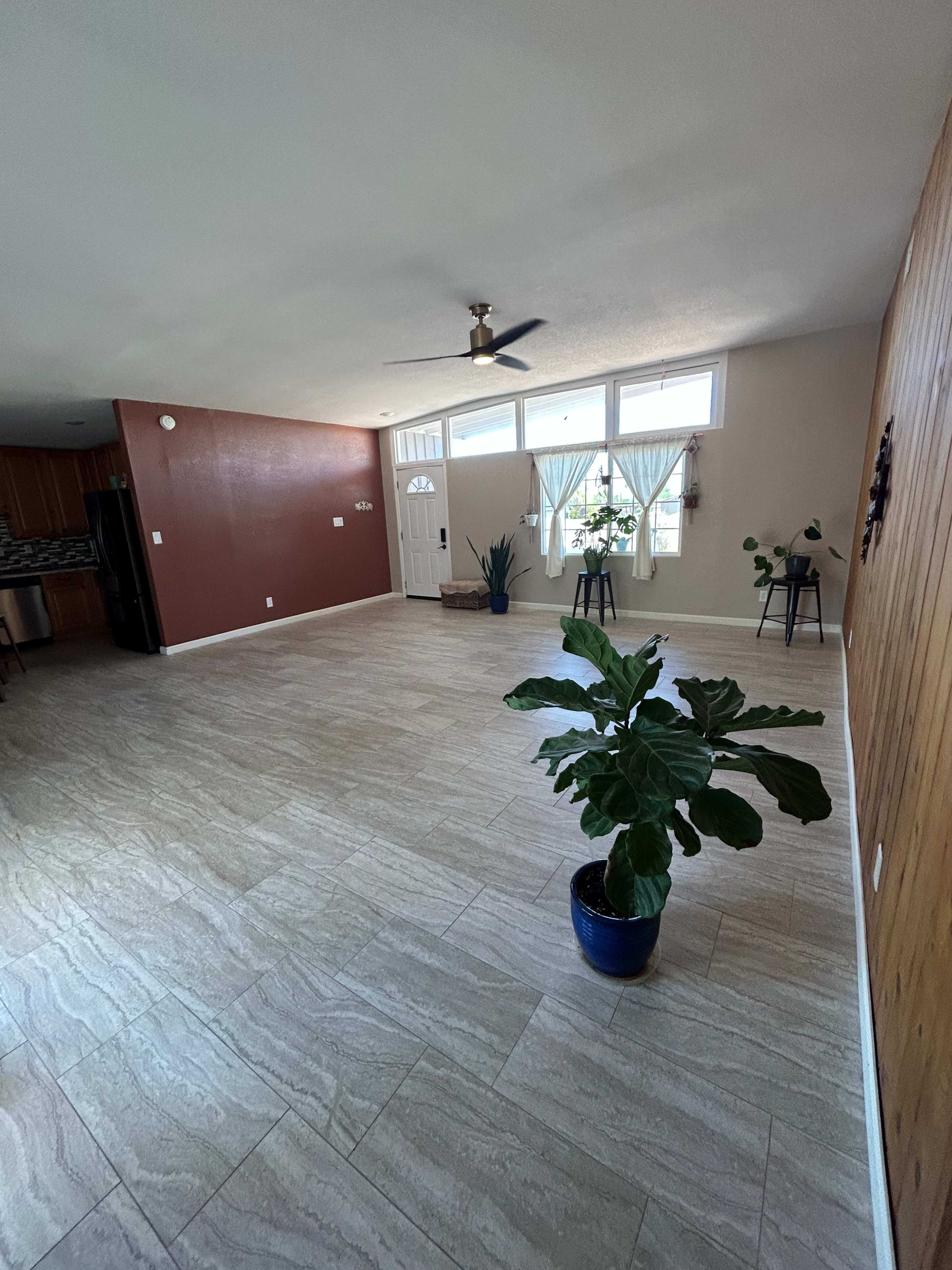 The image shows a spacious living room with tile flooring, a ceiling fan, and a potted plant in the center, featuring large windows and a door in the background.