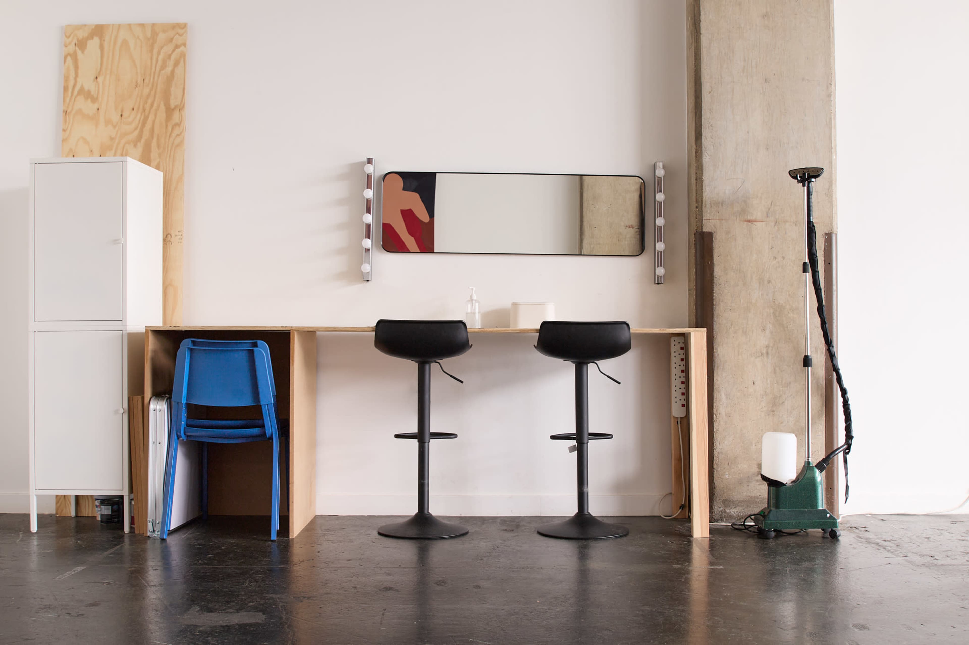 A minimalist workspace features two black stools at a wooden desk with a large mirror overhead and a tall cleaning device nearby.