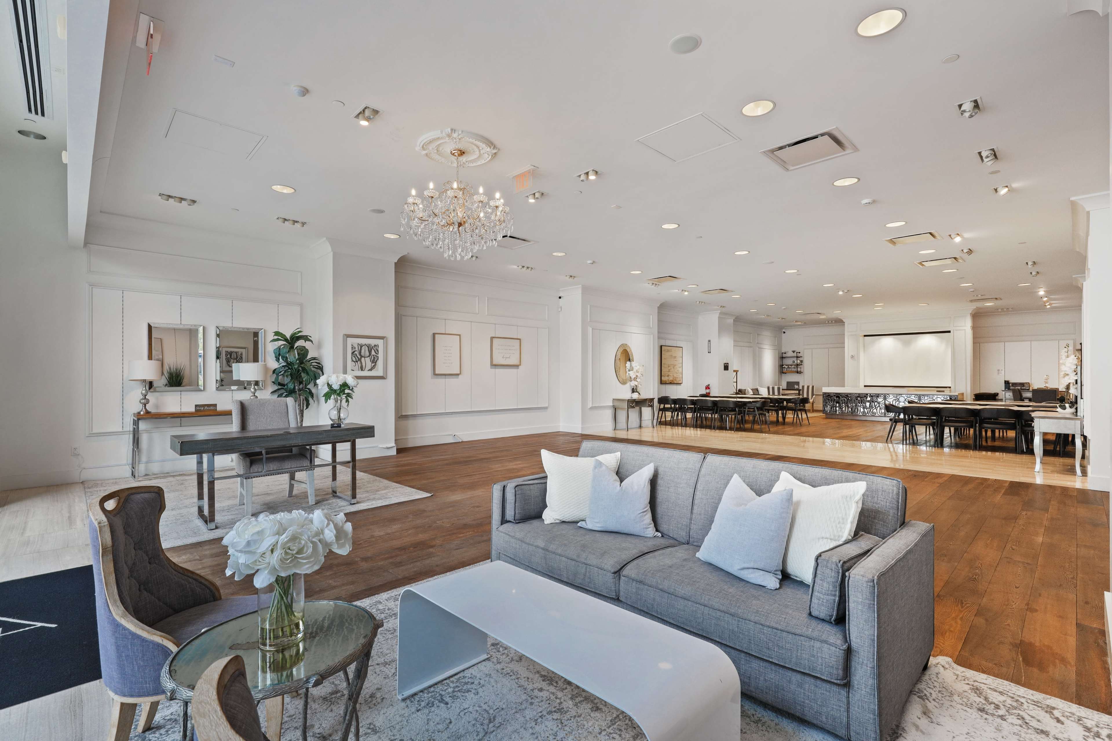 The image shows a brightly lit modern interior space featuring a living area with a gray sofa and coffee table, adjacent to a dining area with multiple tables and chairs.