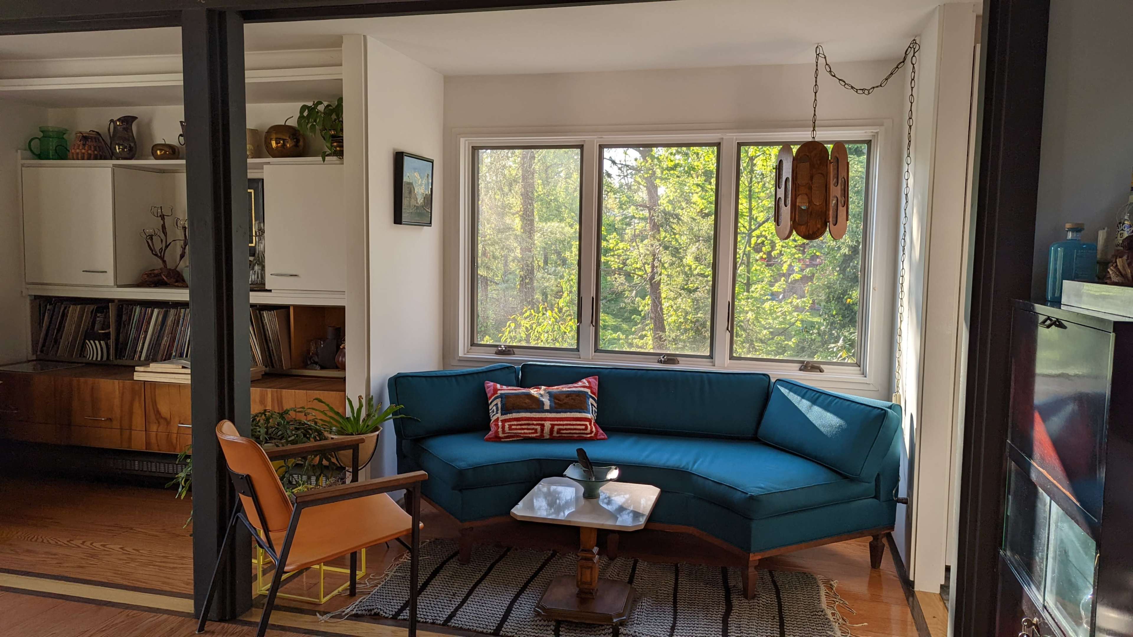 A bright living room features a teal sectional sofa, a wooden coffee table, and large windows overlooking lush greenery.