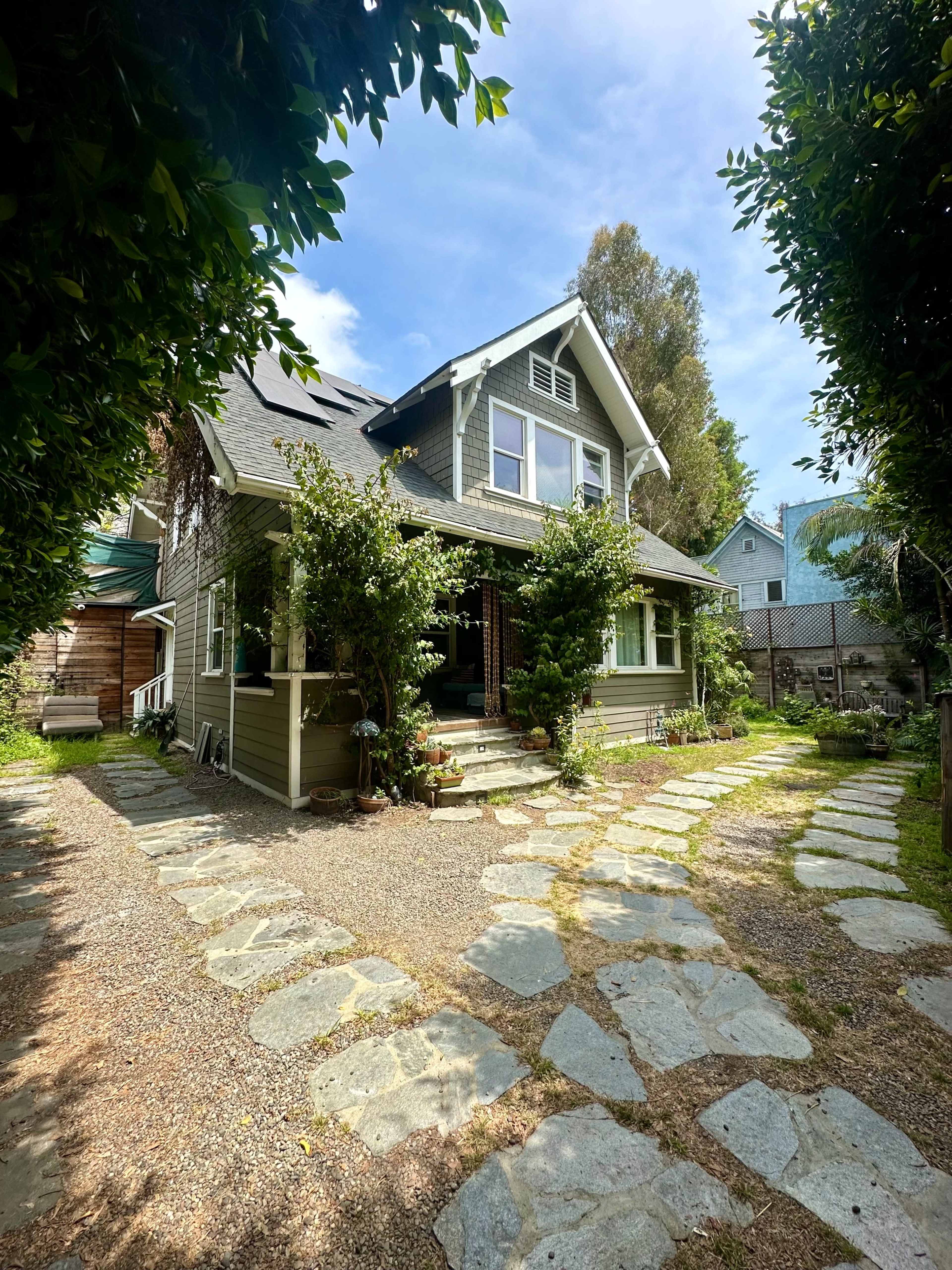 A two-story house with a gabled roof is surrounded by greenery and stone pathways leading to the entrance.