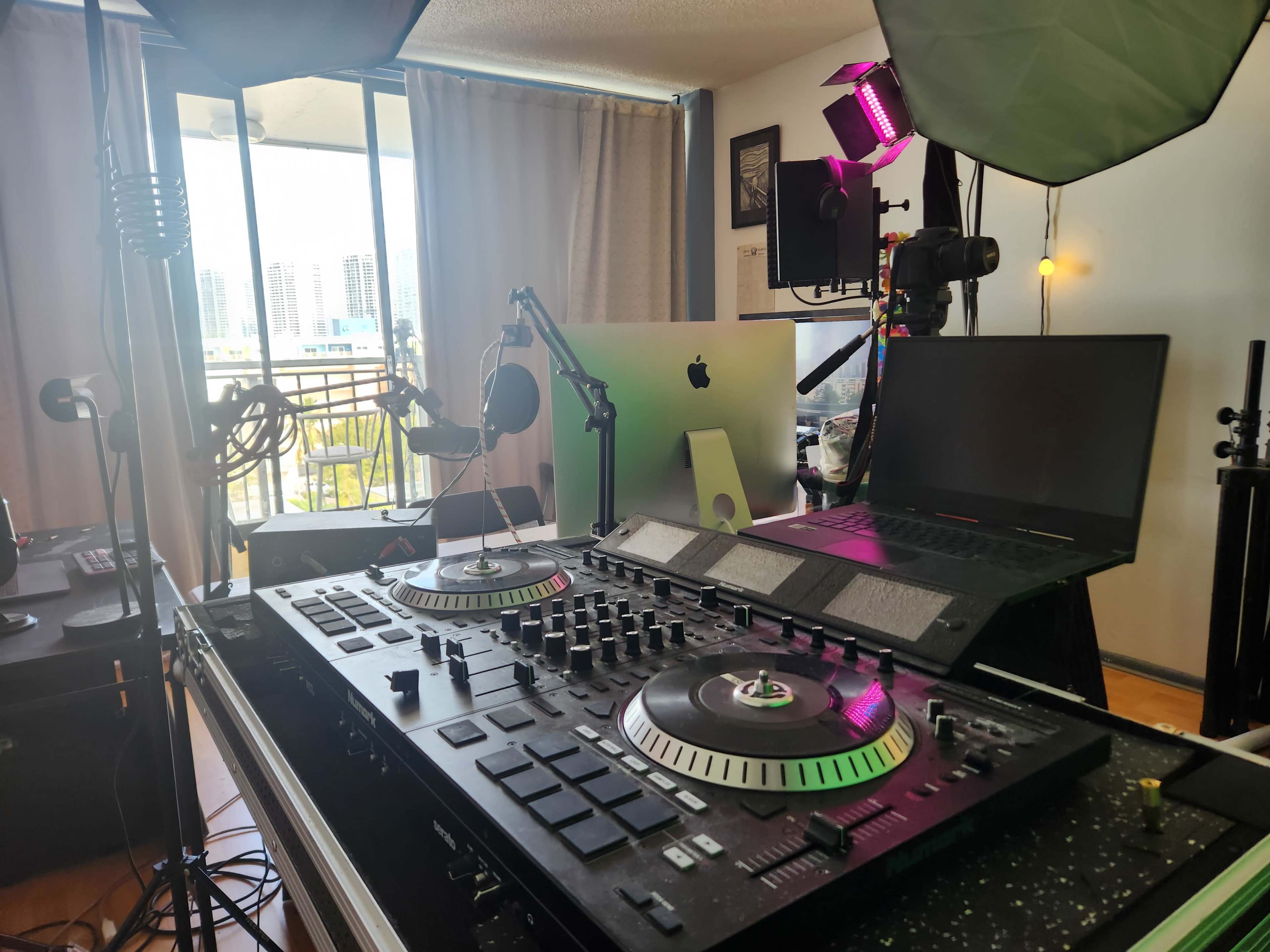 The image shows a DJ setup featuring turntables, a laptop, and studio equipment in a room with a large window and city view.
