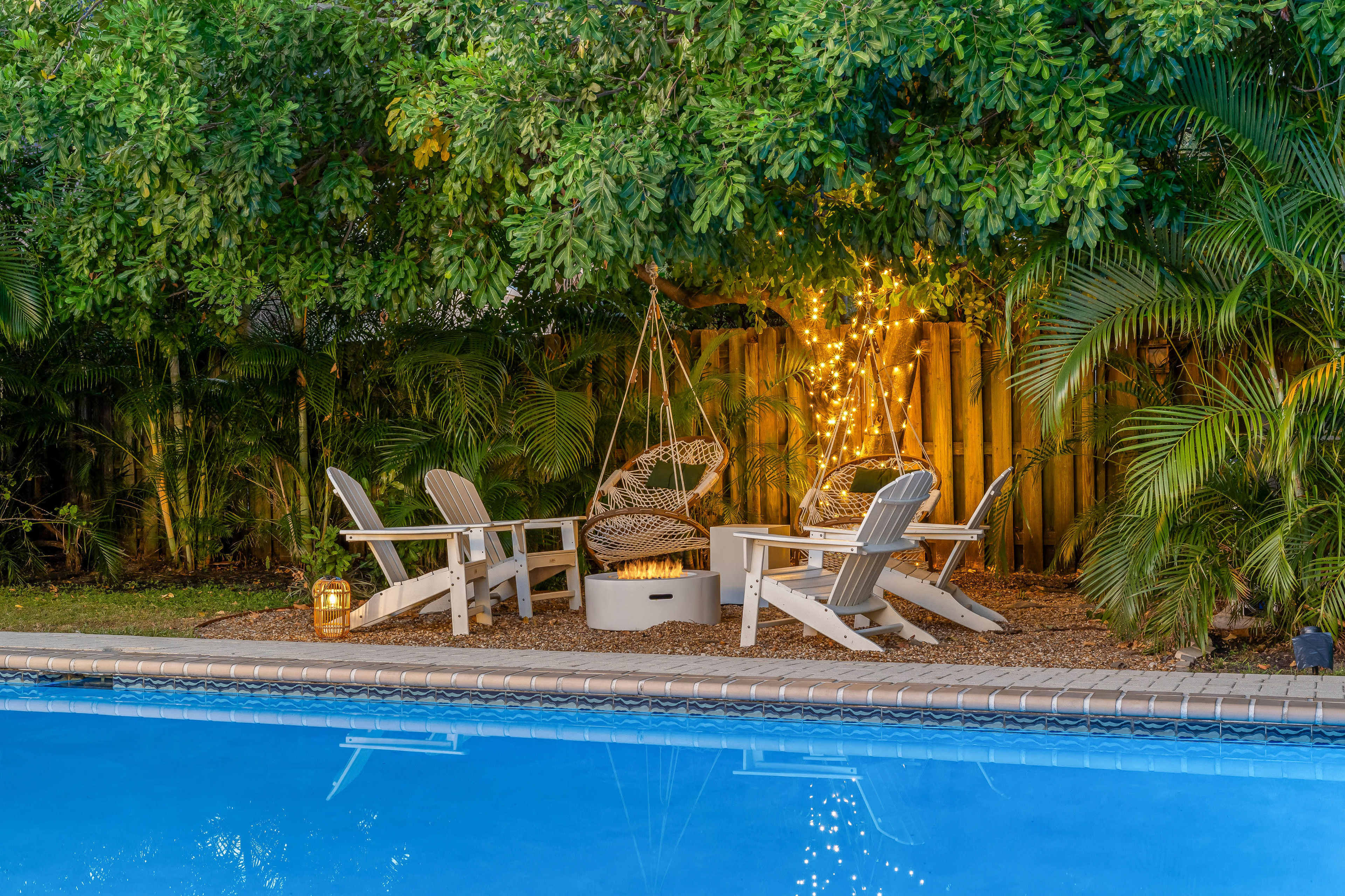 A group of white Adirondack chairs surrounds a circular table near a swimming pool, with a hanging chair and string lights in the background.