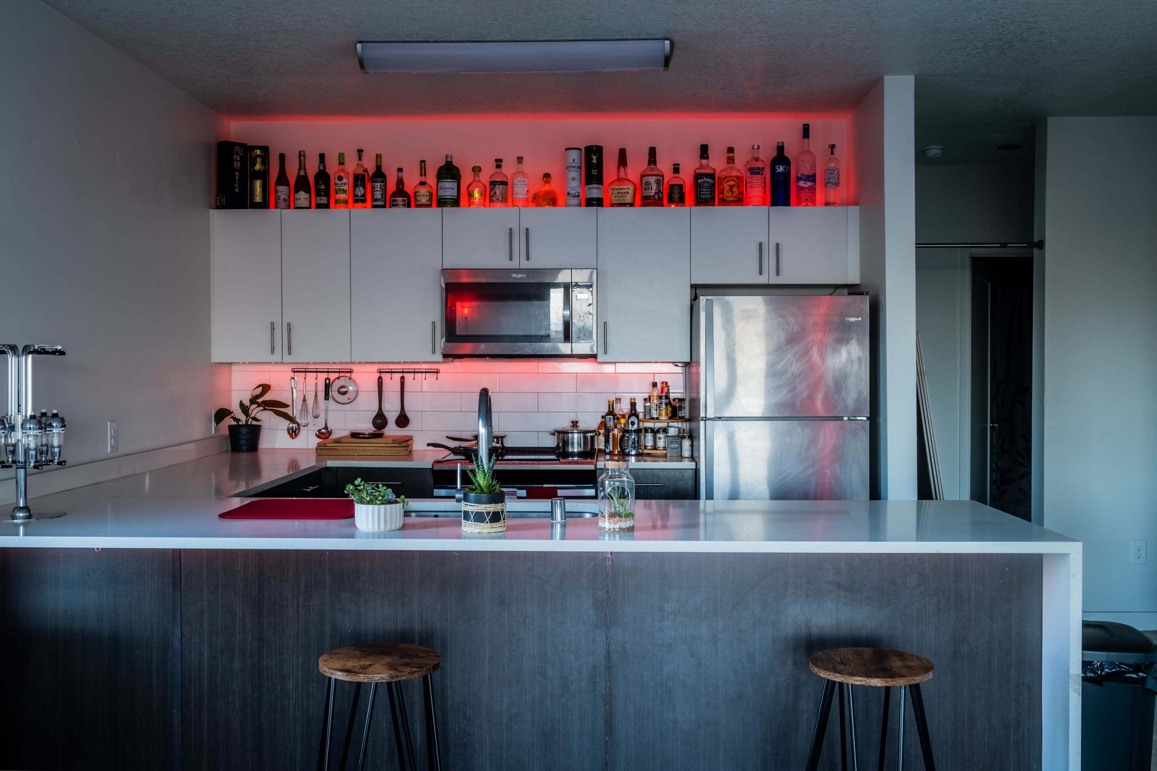 The kitchen features a modern design with a white countertop, stainless steel appliances, and a row of bottles illuminated by red lighting along the upper cabinets.