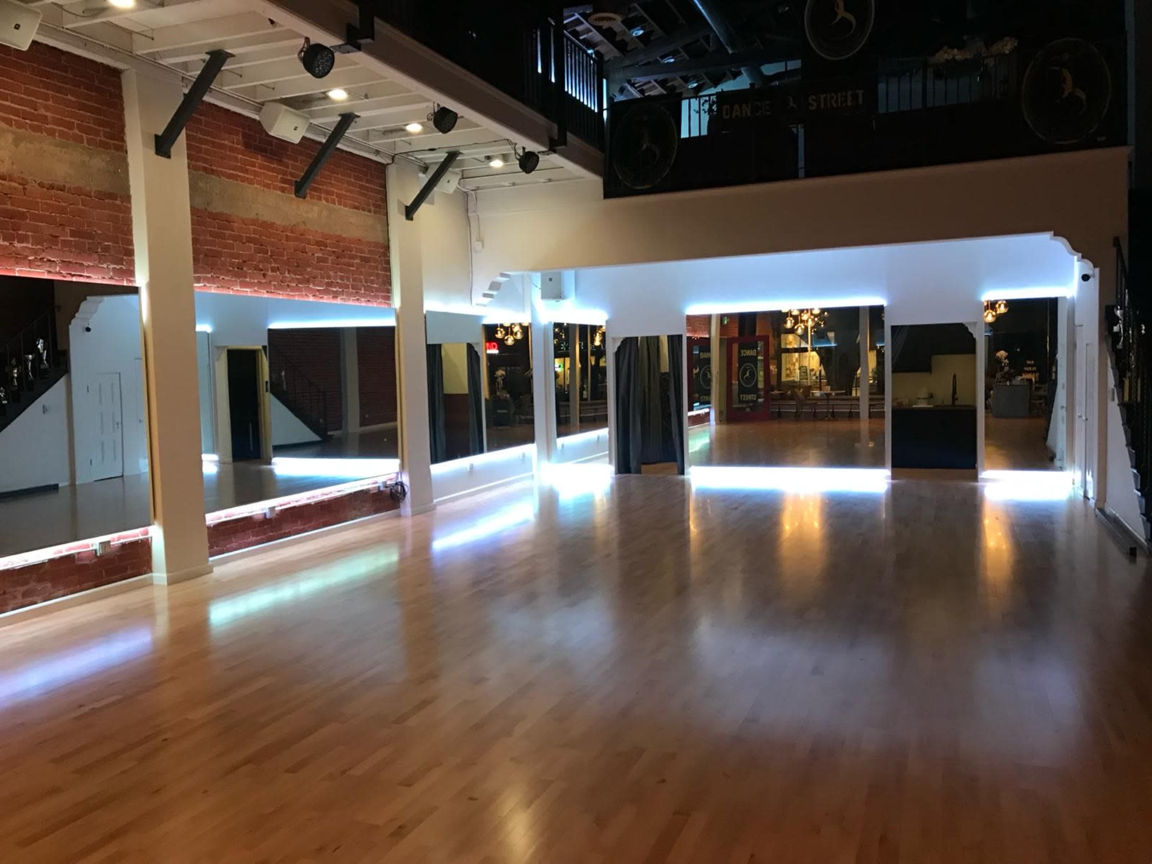 The image shows a spacious dance studio with polished wooden floors, exposed brick walls, and illuminated mirrors along the perimeter.