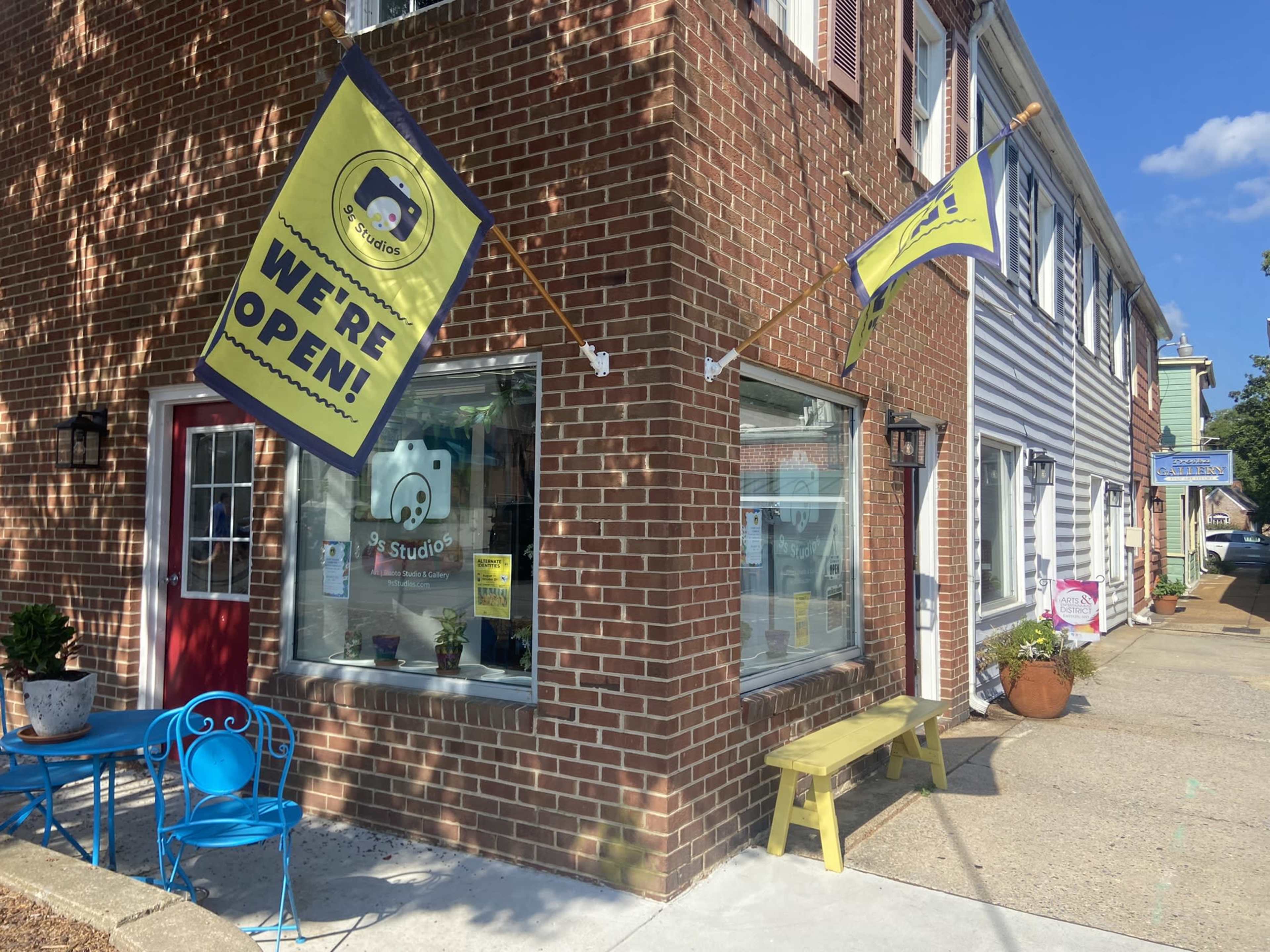 The image shows a brick building with hanging banners that read "WE'RE OPEN," featuring a colorful entrance and outdoor seating with blue and yellow furniture.