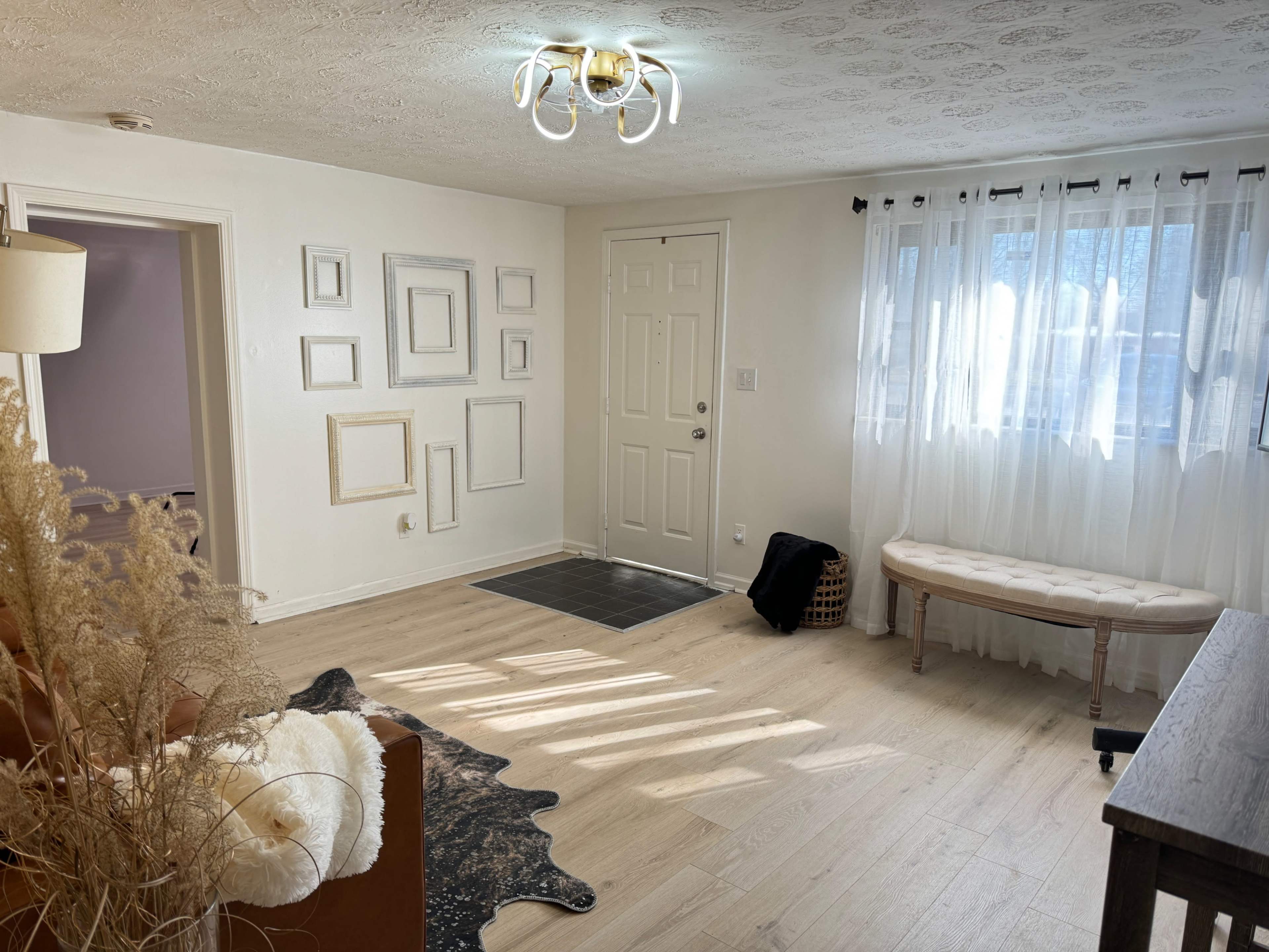 A well-lit entryway with a decorative wall featuring empty frames, a bench, and a window with sheer curtains.