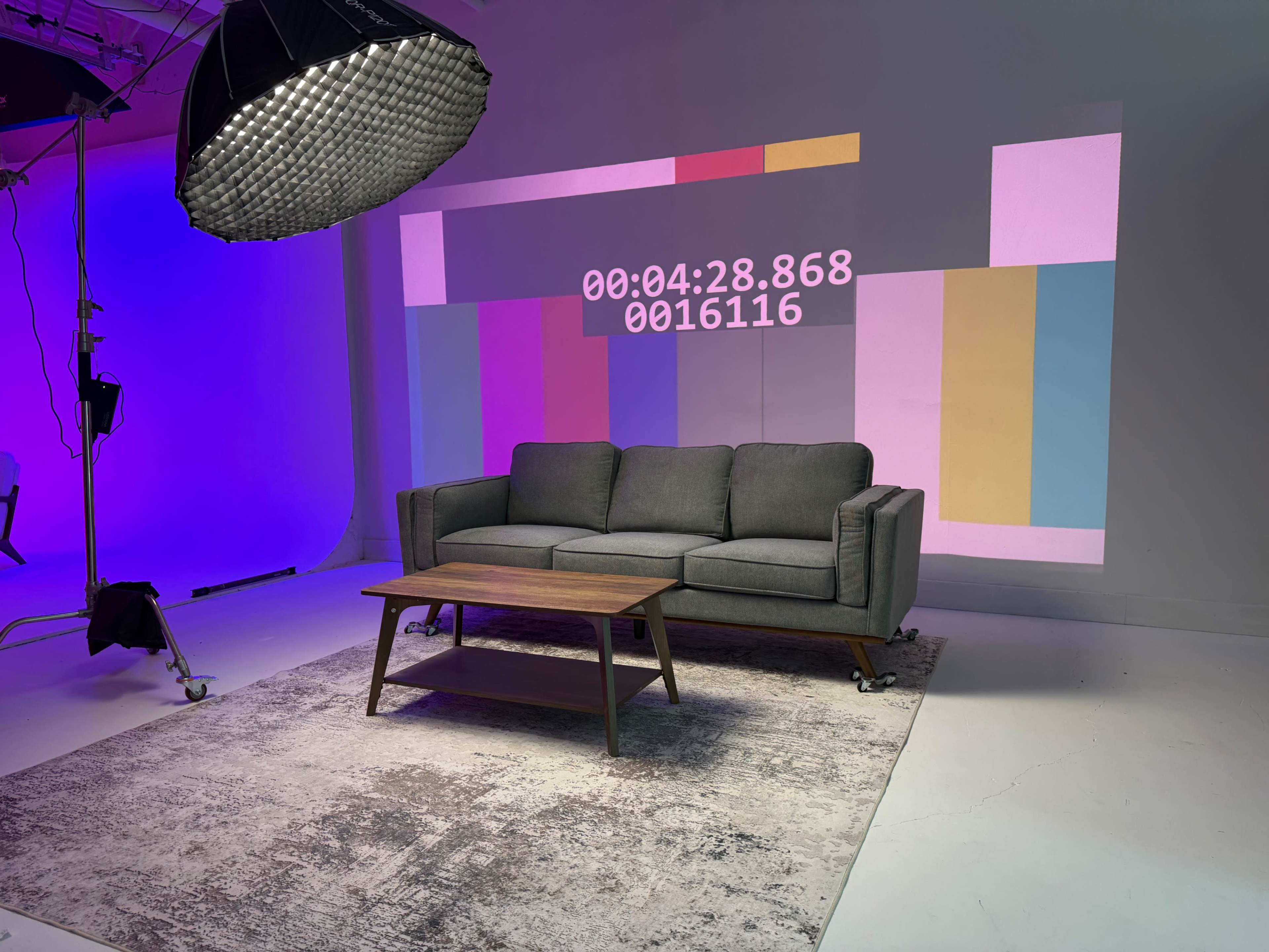 The image shows a gray sofa with a wooden coffee table in front of it, set against a wall displaying colorful geometric projections and a digital timer.