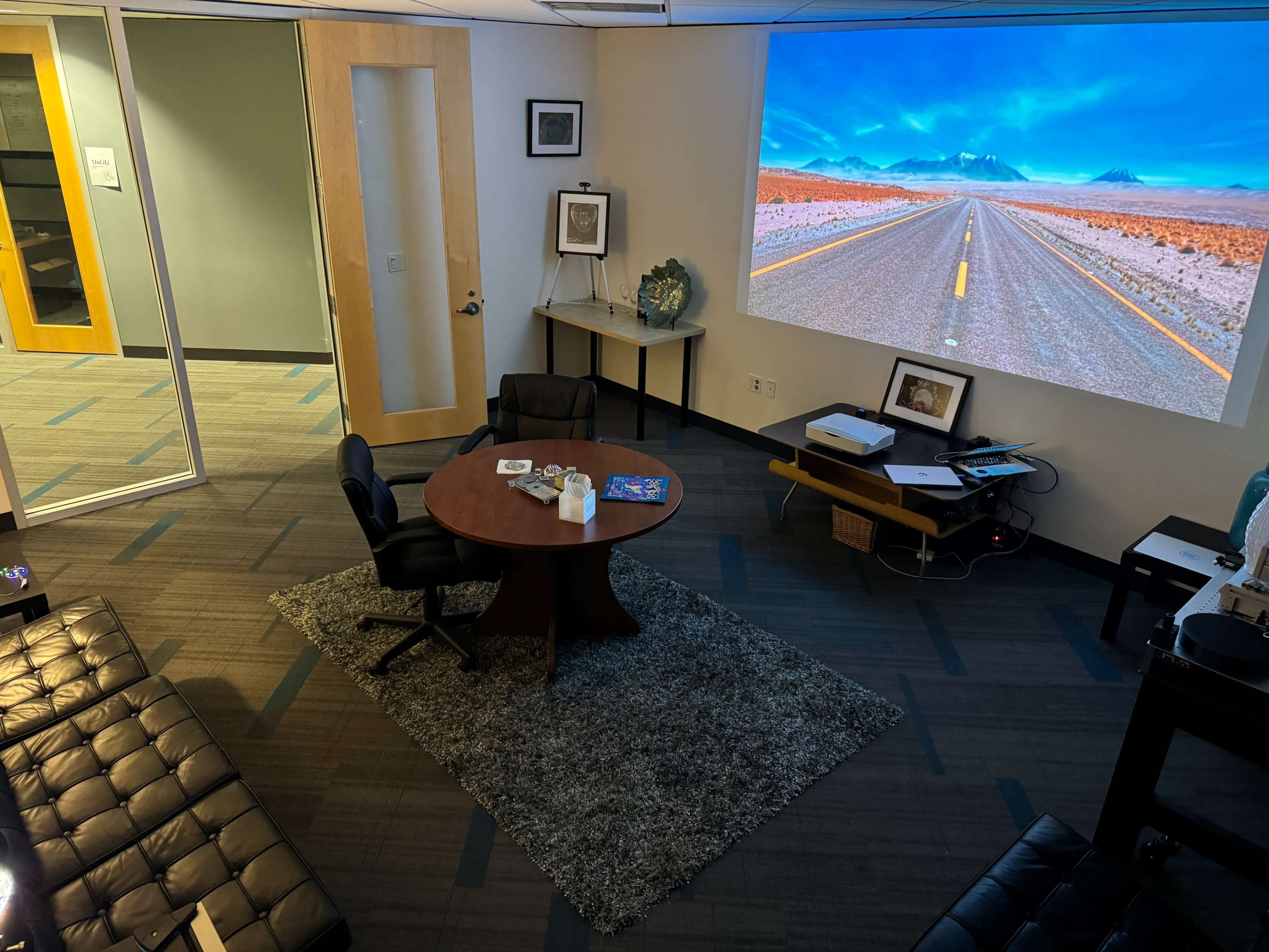 A modern office space features a round table with chairs, a large projection of a road scene on the wall, and minimalist furnishings.