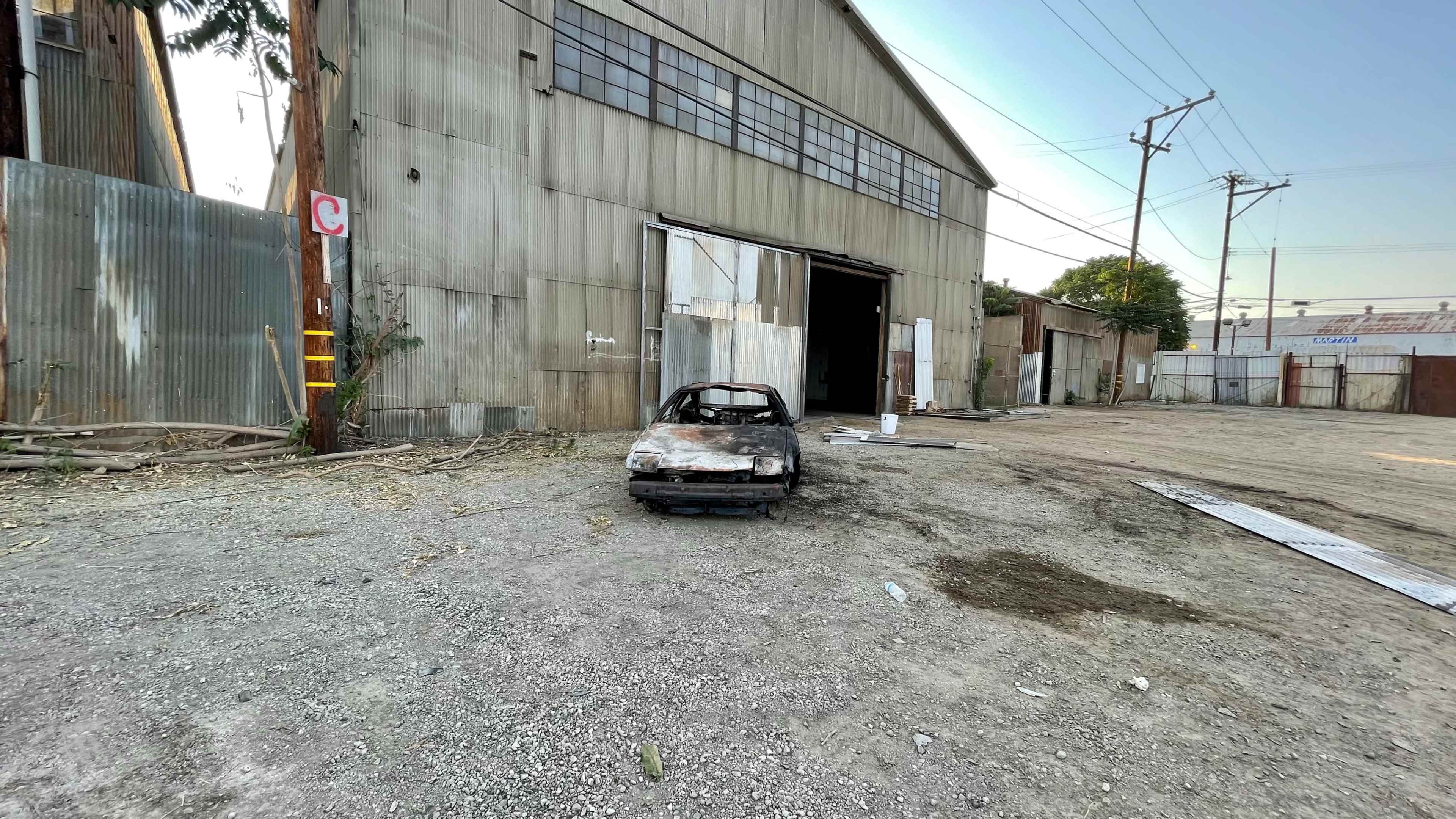 A charred car is parked in front of a large, weathered metal building on a gravel lot.