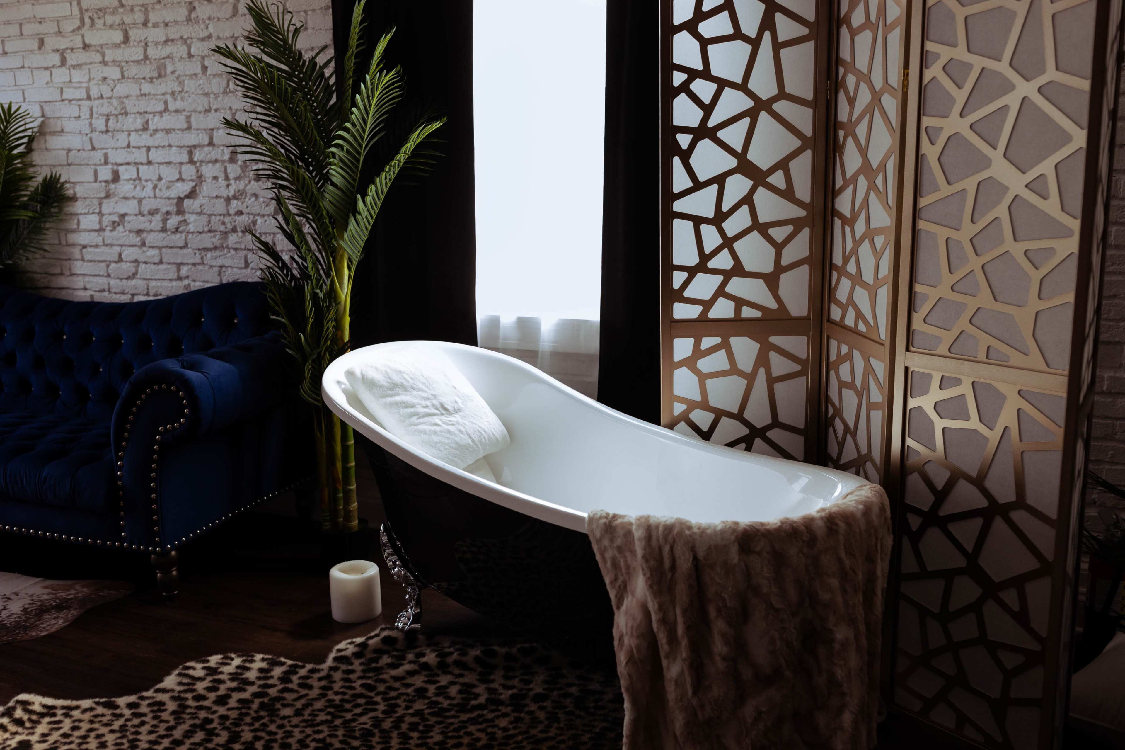 A freestanding bathtub is positioned next to a chaise lounge in a room with decorative wood screens and a patterned rug.