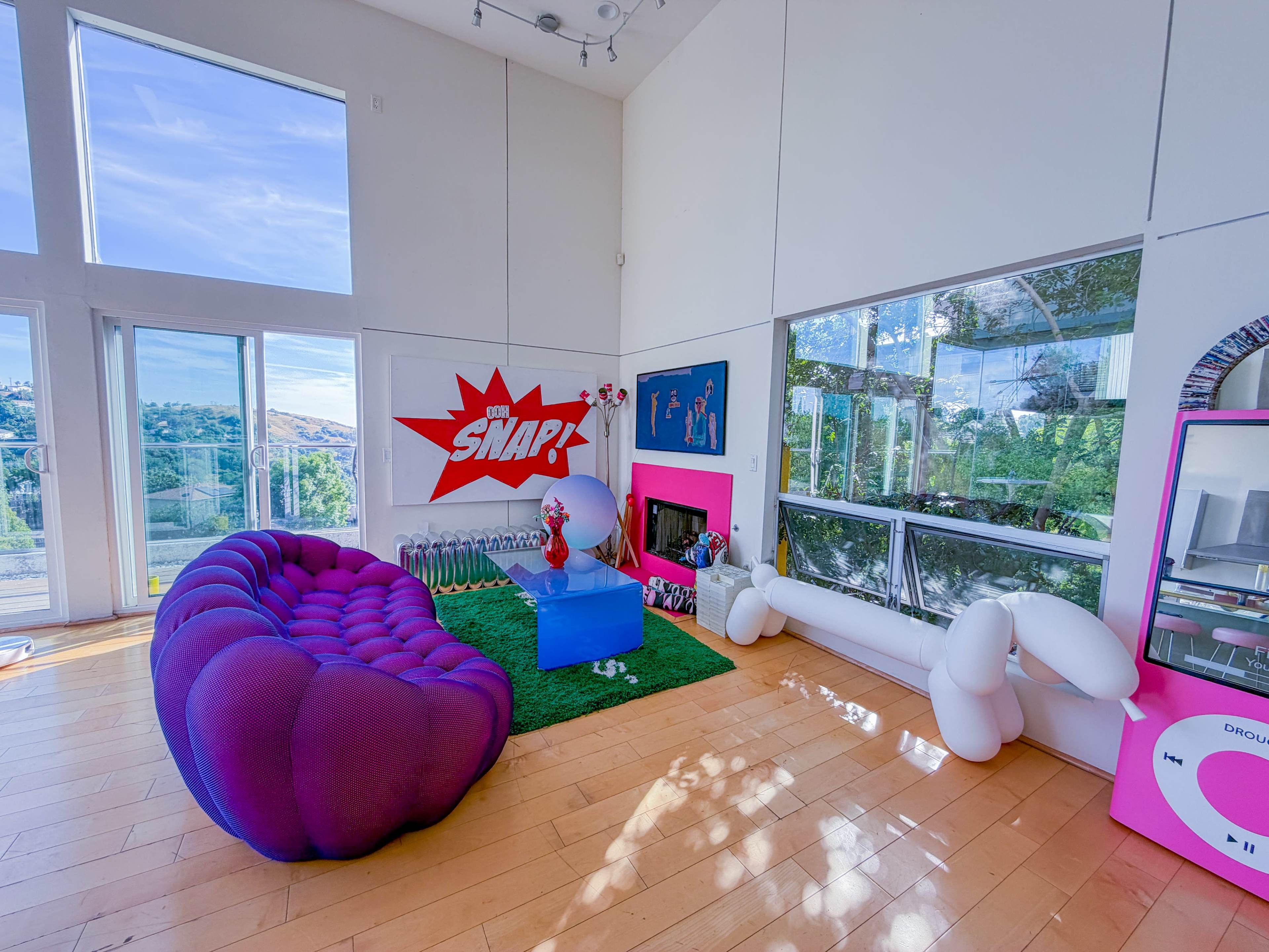 The image shows a modern living room with large windows, featuring a purple sofa, a green rug, and colorful wall decor.