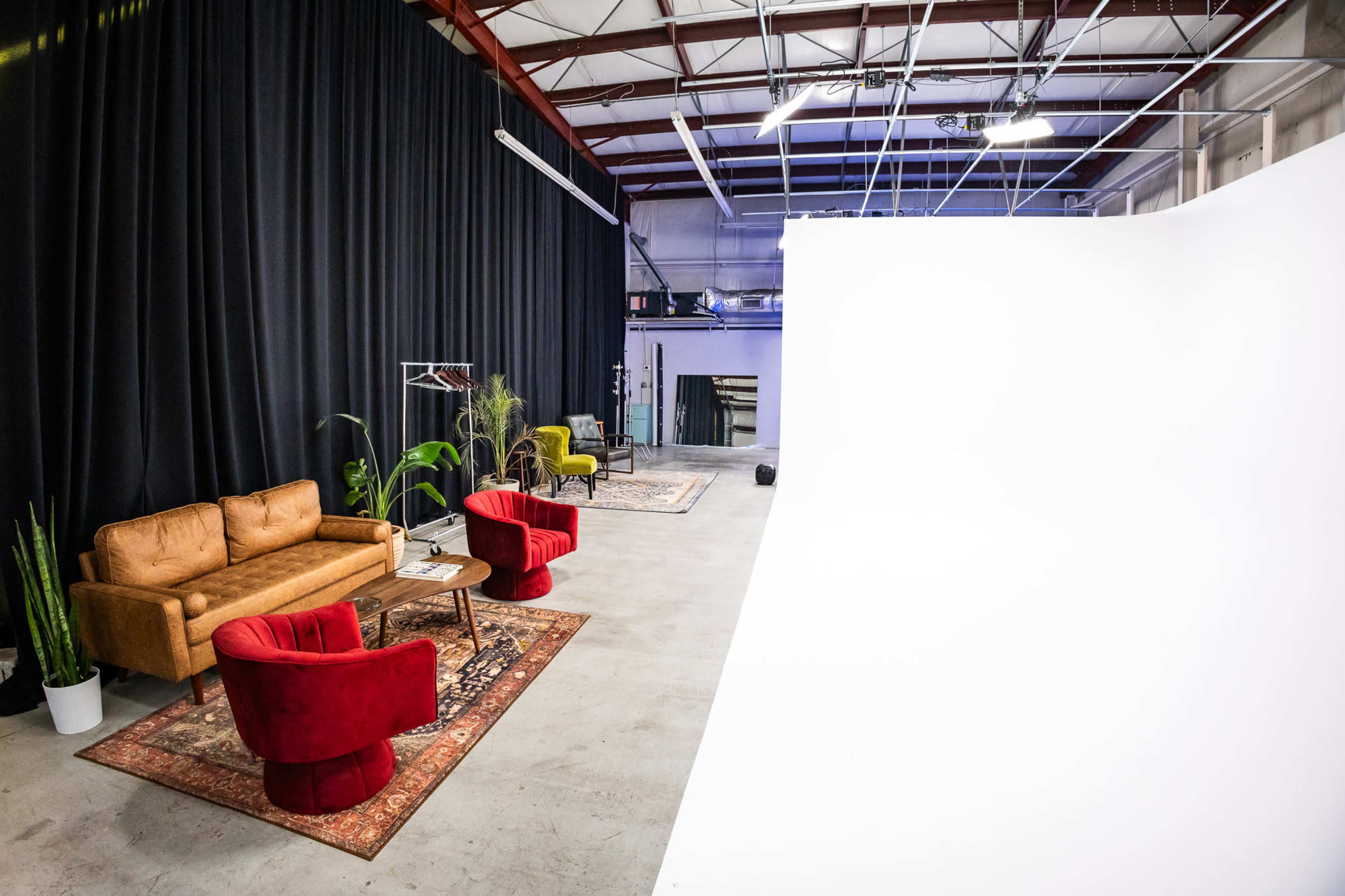 The image shows a spacious studio with a white backdrop, two red chairs, a brown couch, a small table, and various plants, all arranged on a gray floor.