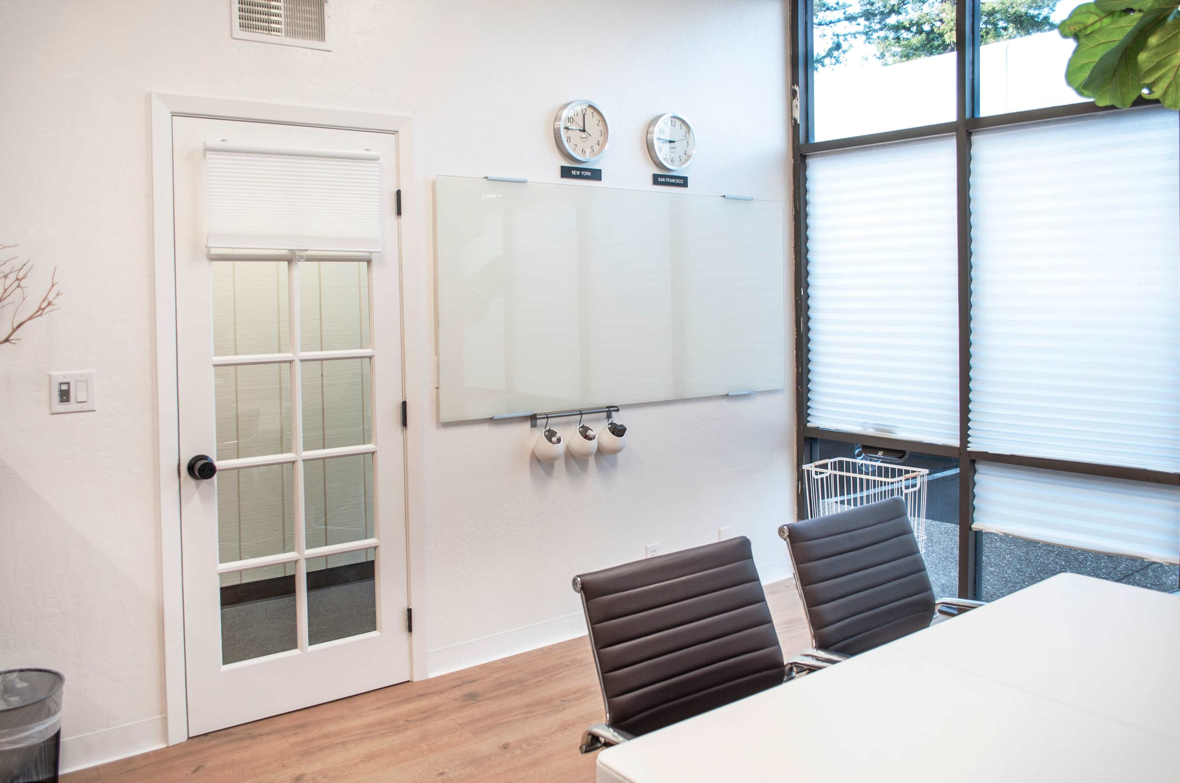 A modern office space with a glass door, a whiteboard mounted on the wall, two wall clocks, and a table surrounded by black chairs.