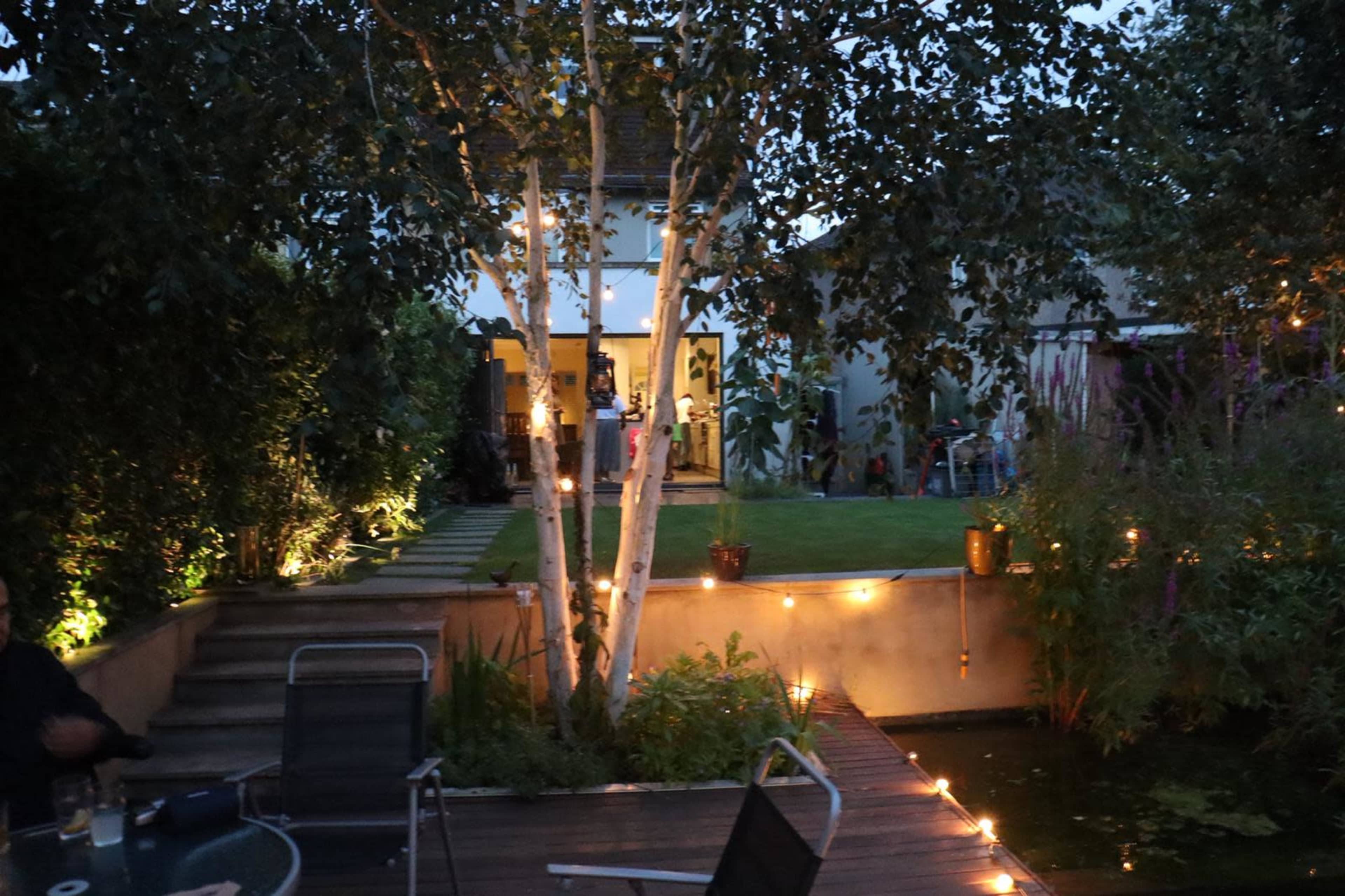A well-lit garden features a birch tree, a pond, and a house in the background with an open doorway.