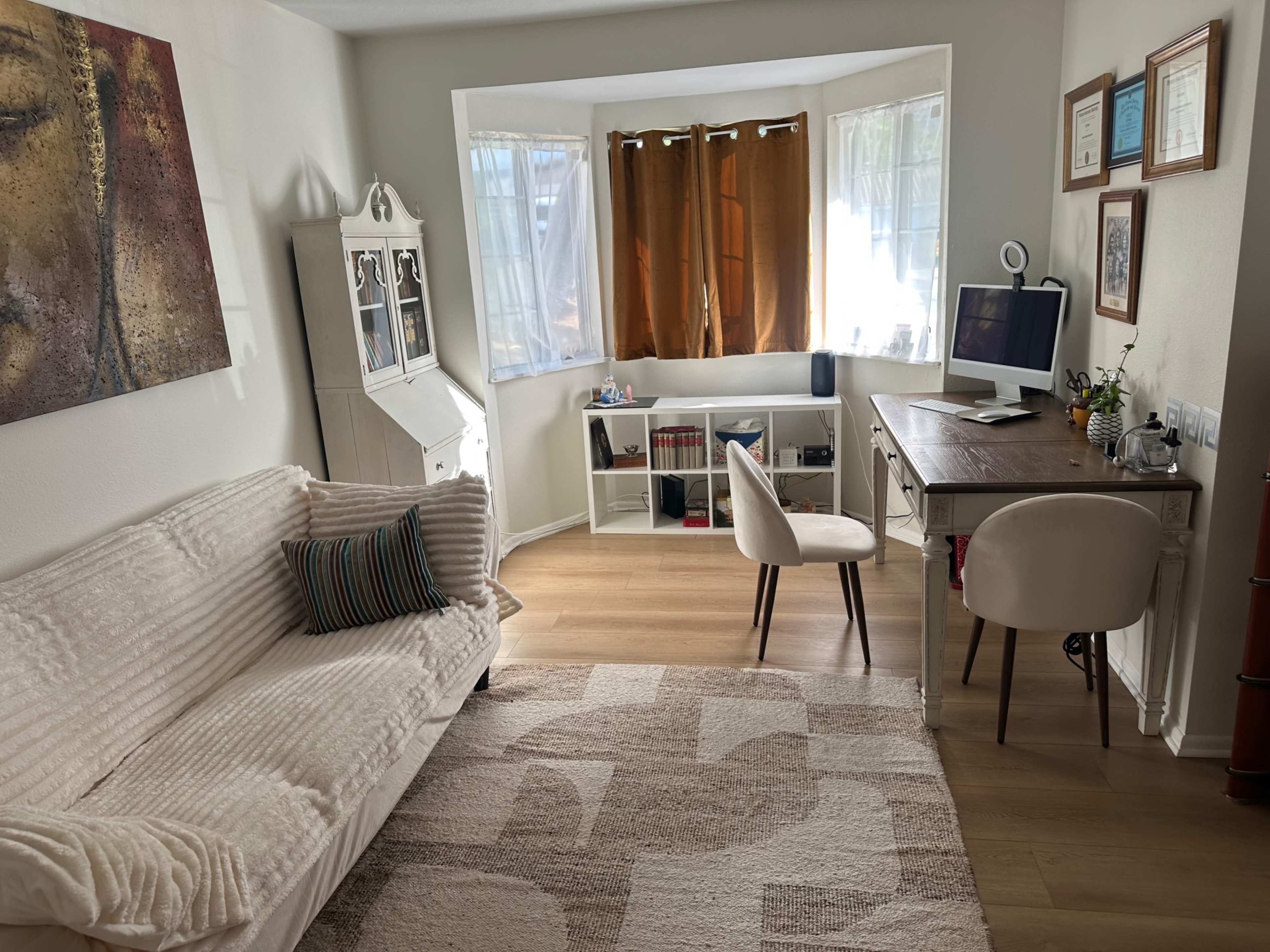 The image shows a cozy room featuring a light-colored sofa, a desk with a chair, a small shelf, and large windows with brown curtains.