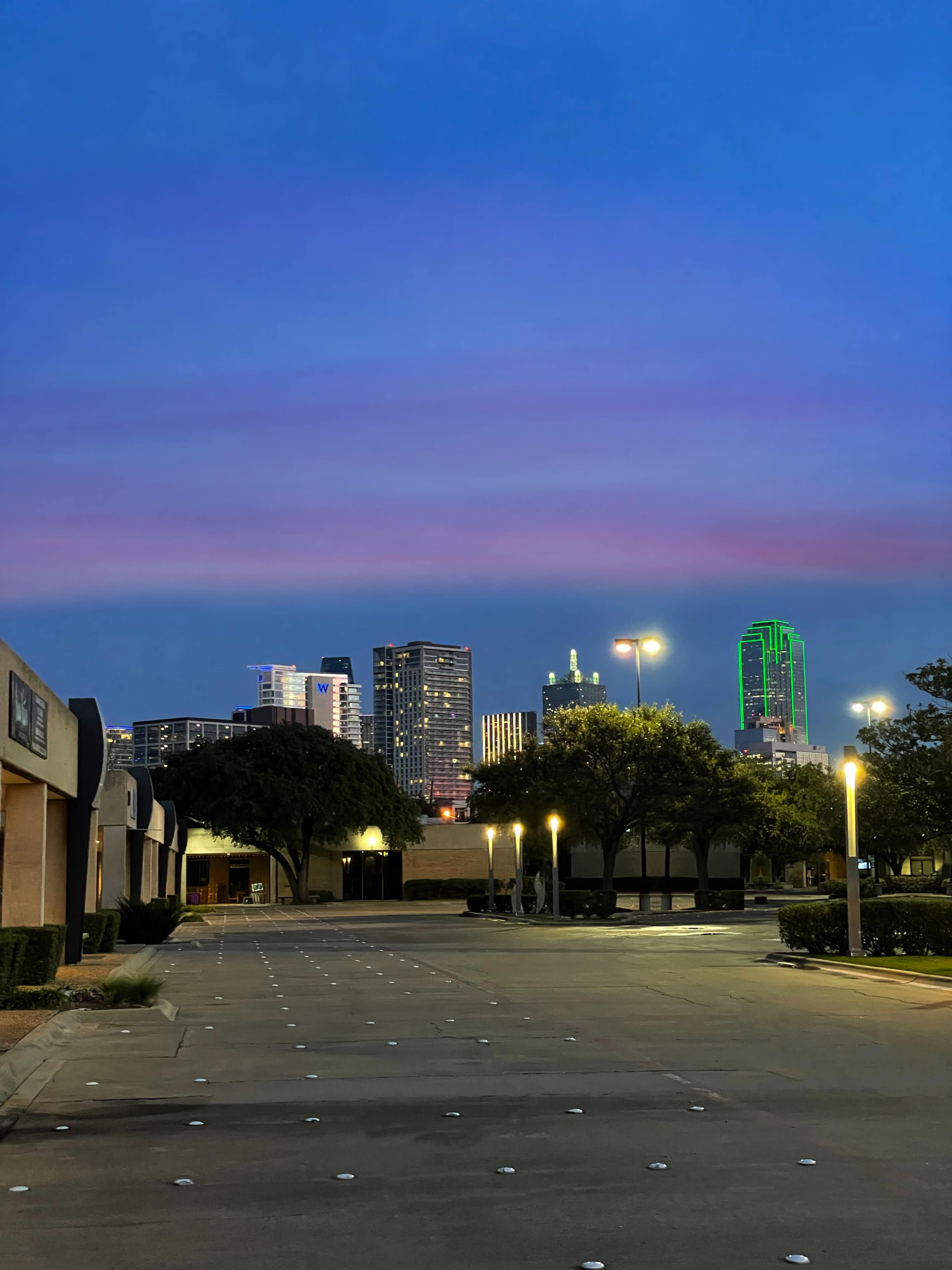 A city skyline with tall buildings illuminated against a twilight sky, viewed from a deserted street lined with trees and streetlights.