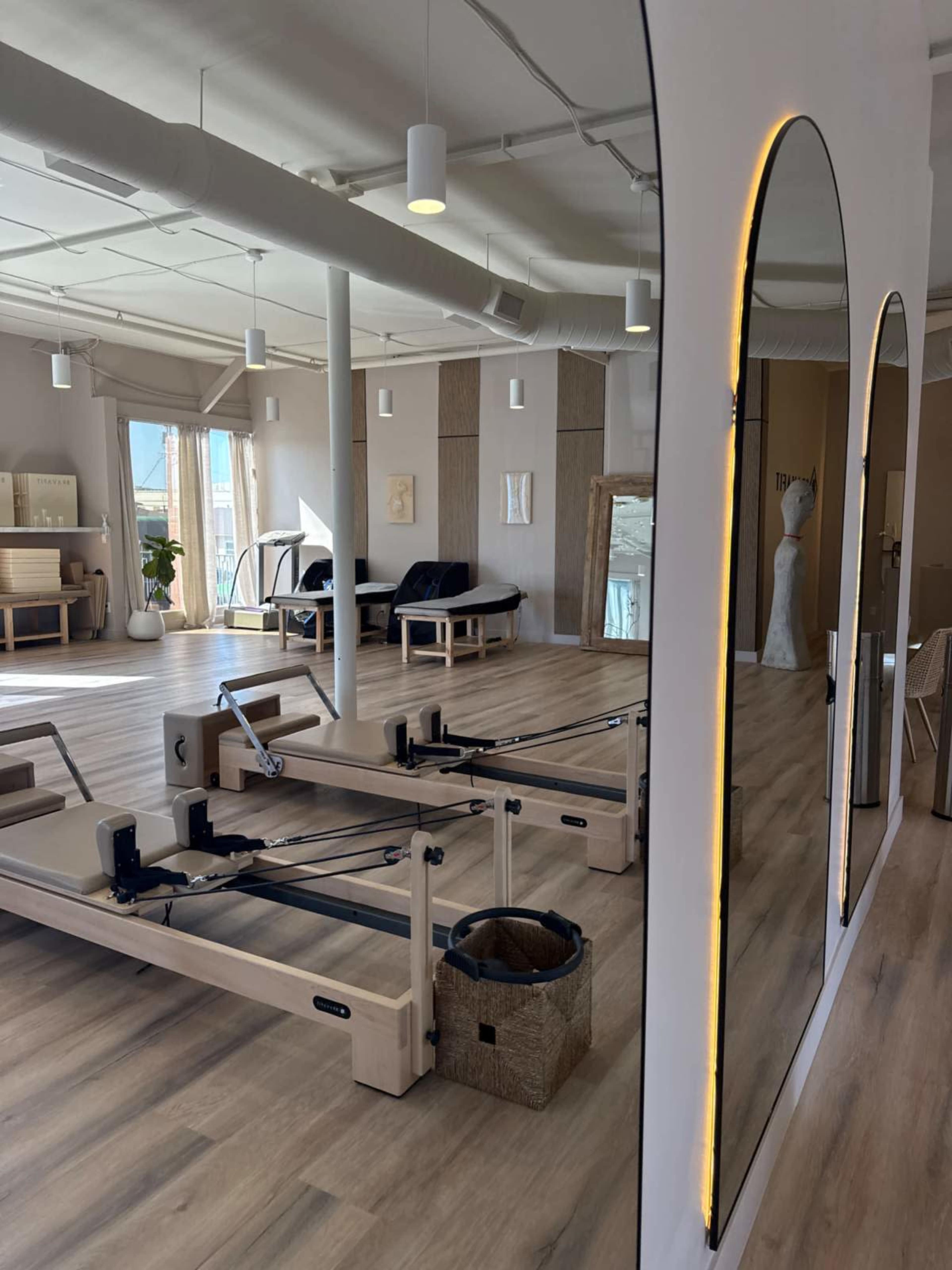 The image shows a spacious fitness studio with wooden floors, several pilates reformers, and large mirrors reflecting the light from windows.