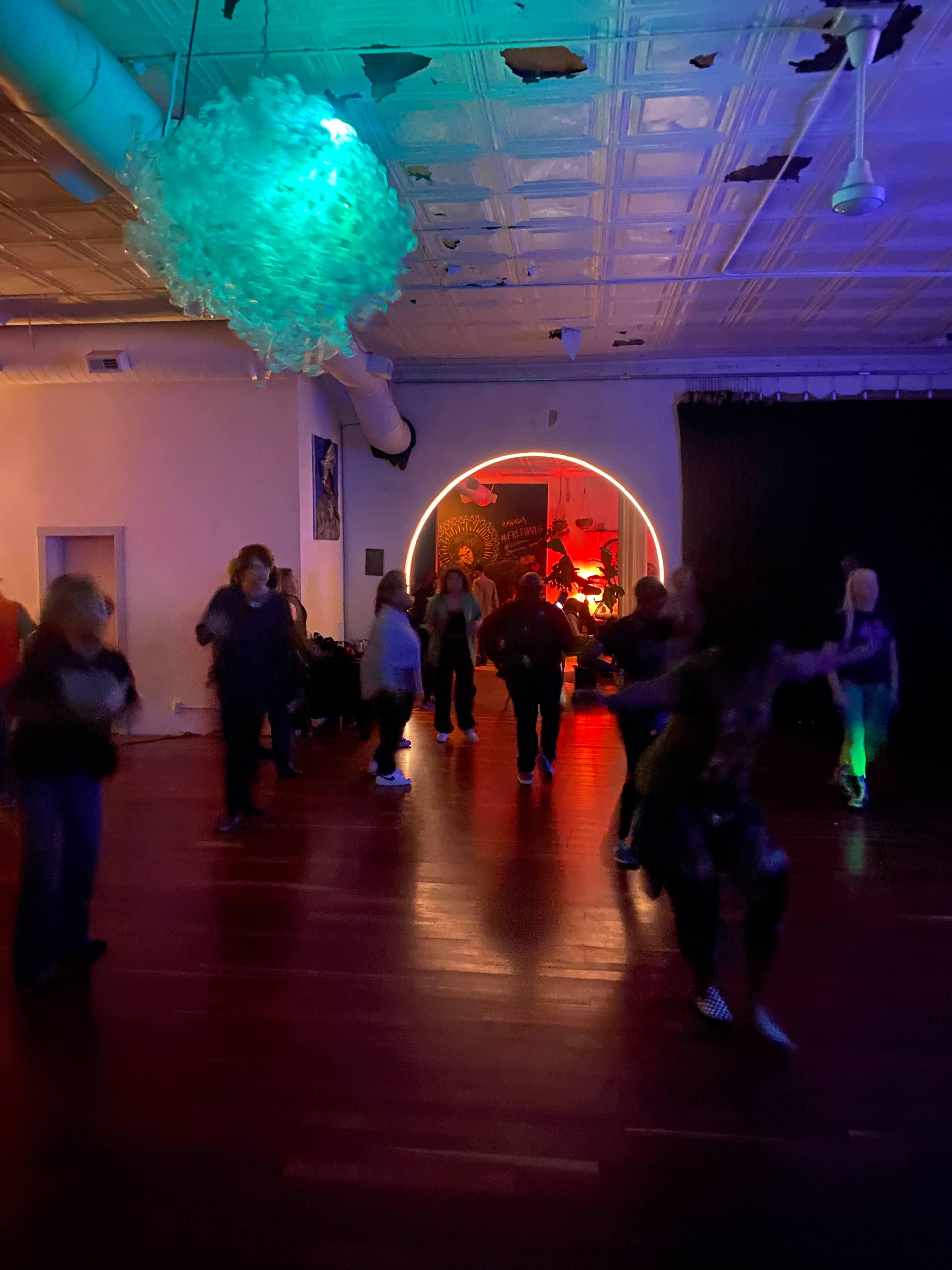 A group of people dance under colorful lights in a room with an illuminated circular backdrop.