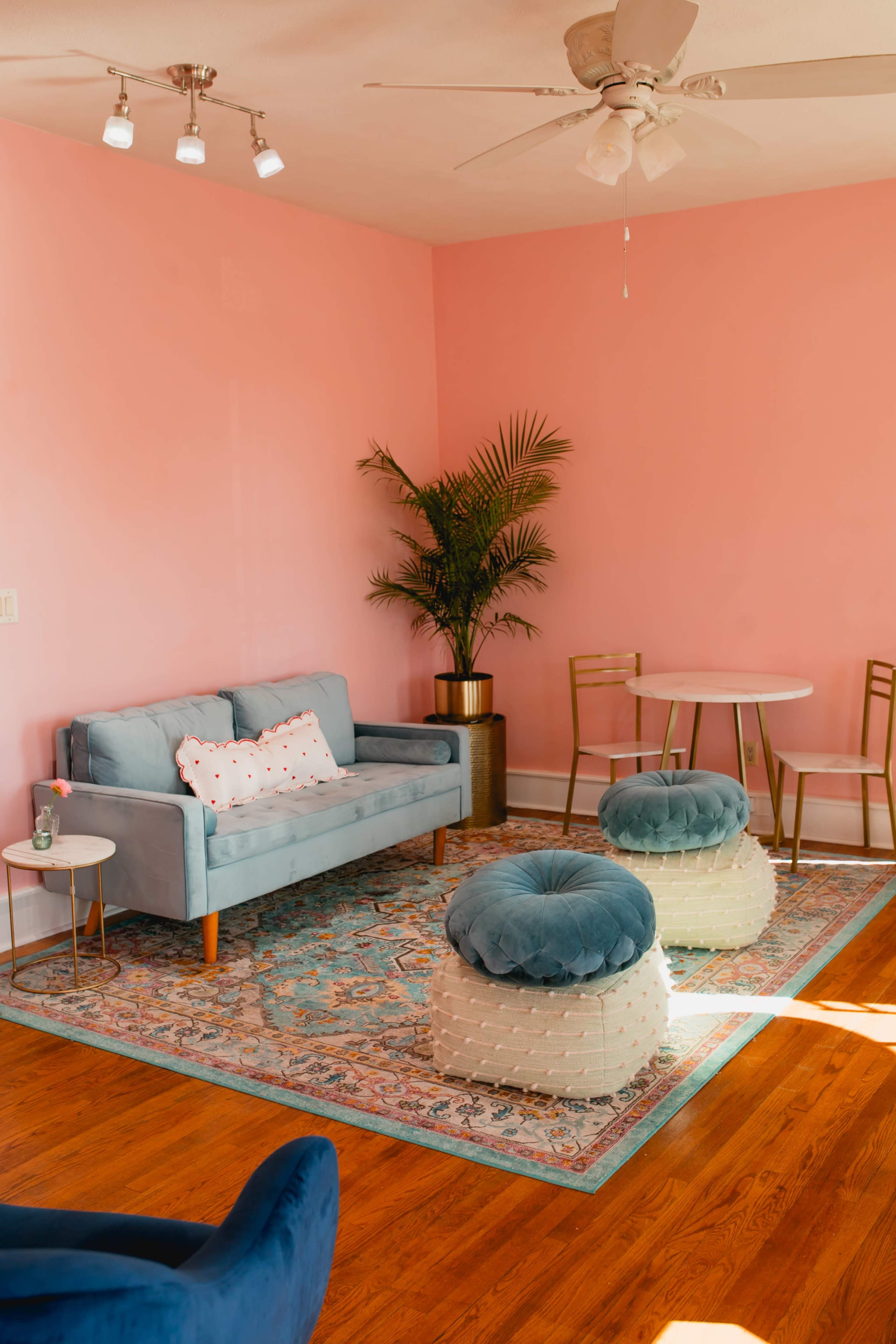A light blue couch with patterned pillows sits on a decorative rug in a room with pink walls, accompanied by a small round dining table and two poufs.