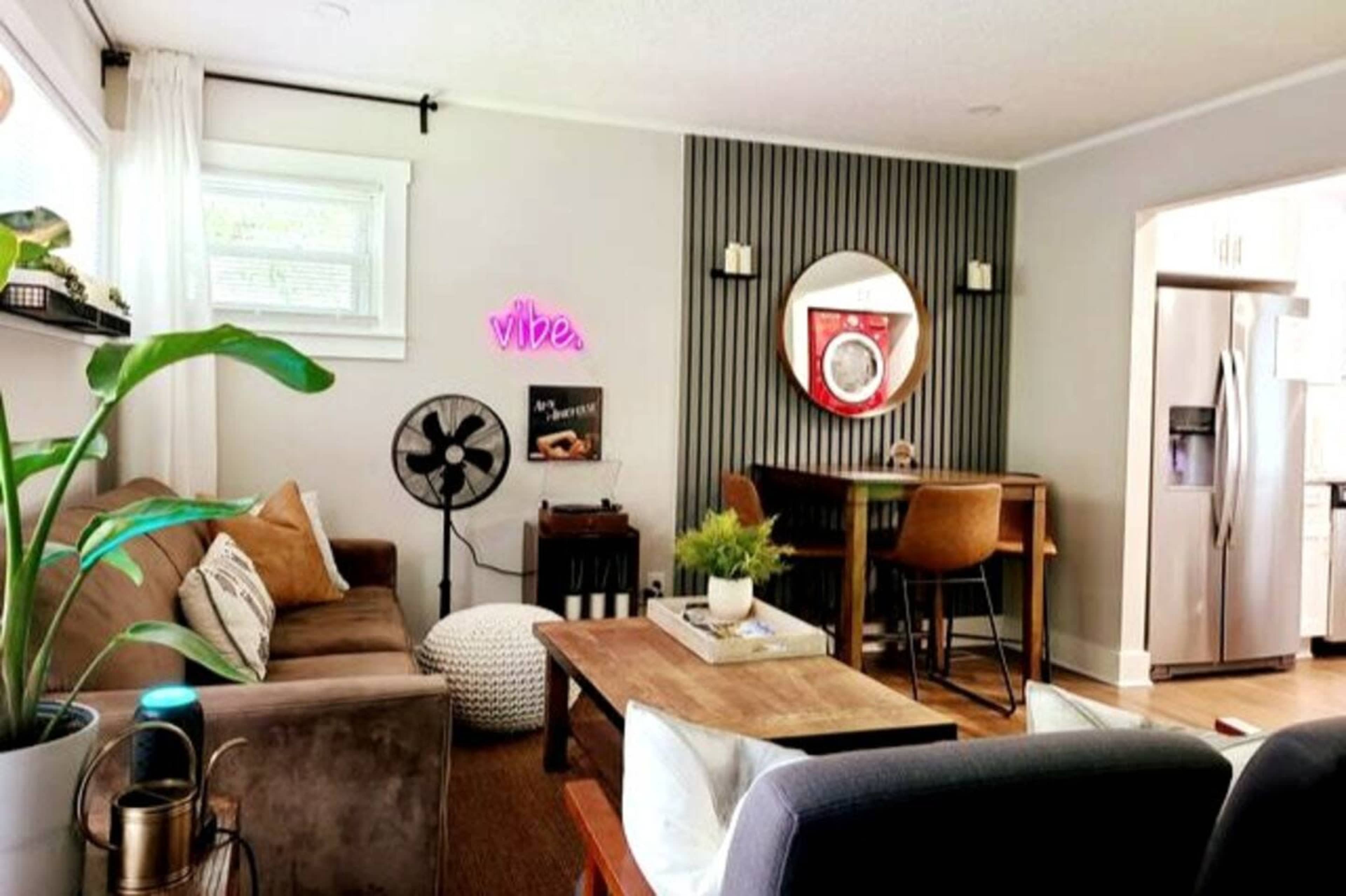 A living room features a brown sofa, a round mirror, a bar-style dining table, and a neon sign that reads "vibe."