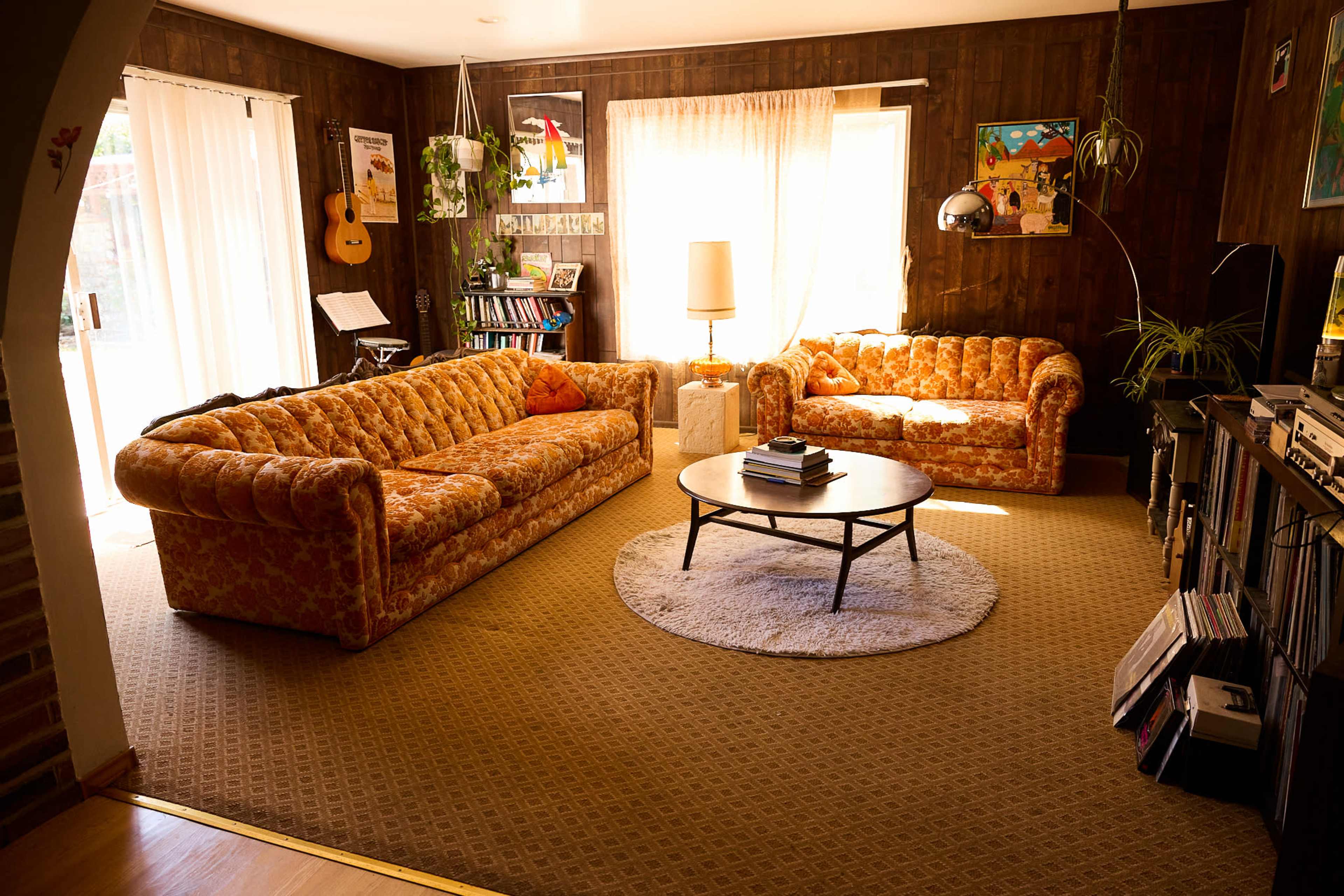 The scene features a living room with orange-patterned sofas, a circular coffee table, and wooden walls, illuminated by natural light from a large window.